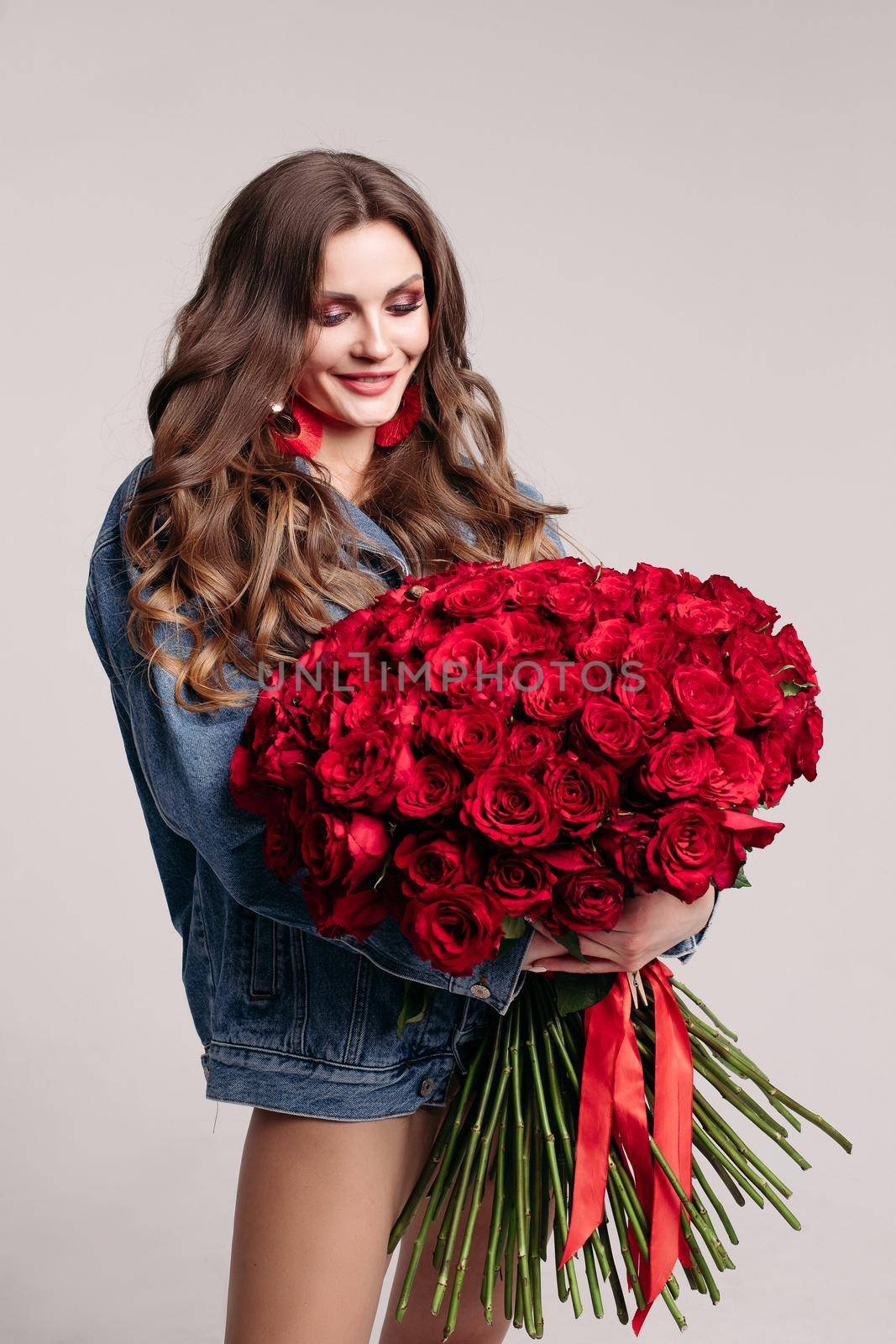 Beautiful brunette girl looking down at huge bouquet of flowers tied with stripes. Elegant lady with long hair standing in jeans jacket. Gorgeous woman with big earings holding roses and smiling.