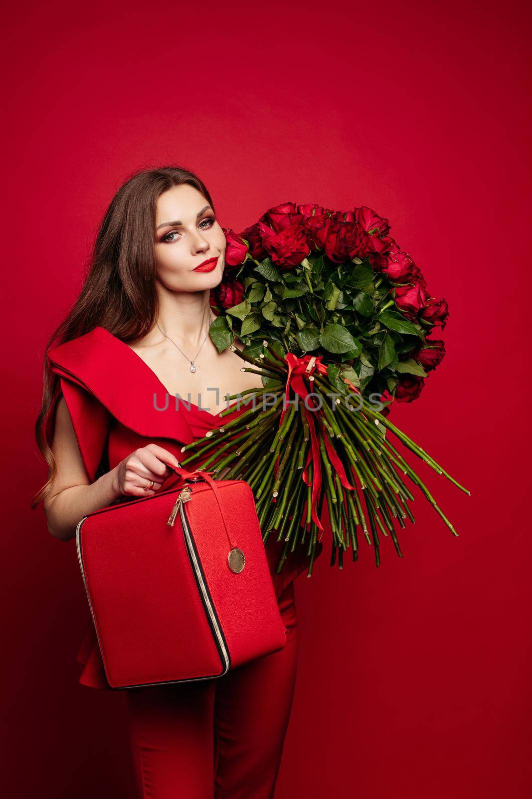 Young woman with elegant jewelry wearing fashionable costume and looking up. Brunette stylish lady in blouse and pants holding bag. Beautiful girl with big bouquet of red roses on shoulder dreaming.
