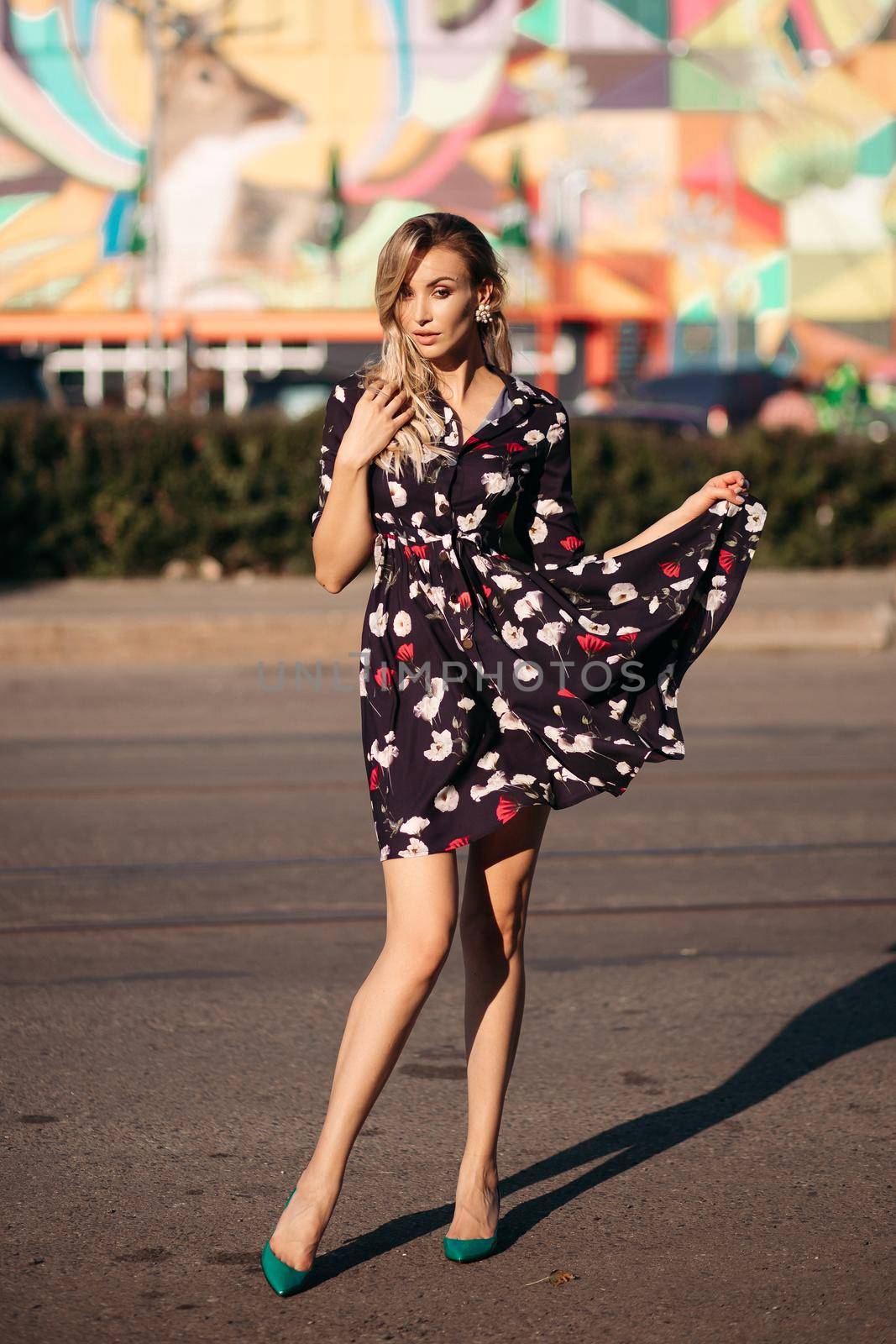 Full length portrait of stunning blonde woman in colored black, white and red dress and green heels posing with skirt in hand and eyes closed in bright sunshine in the street. Colored background.