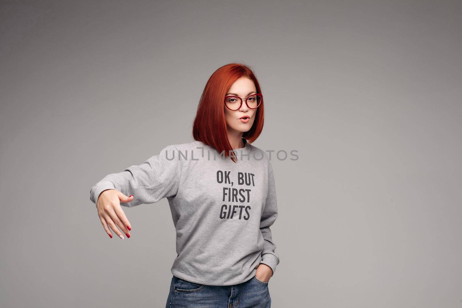 Swag and confident red haired girl in jeans and sweatshirt posing at gray studio, looking at camera and gesturing by hands. Young stylish woman wearing in bag girl look. Concept of fashion.