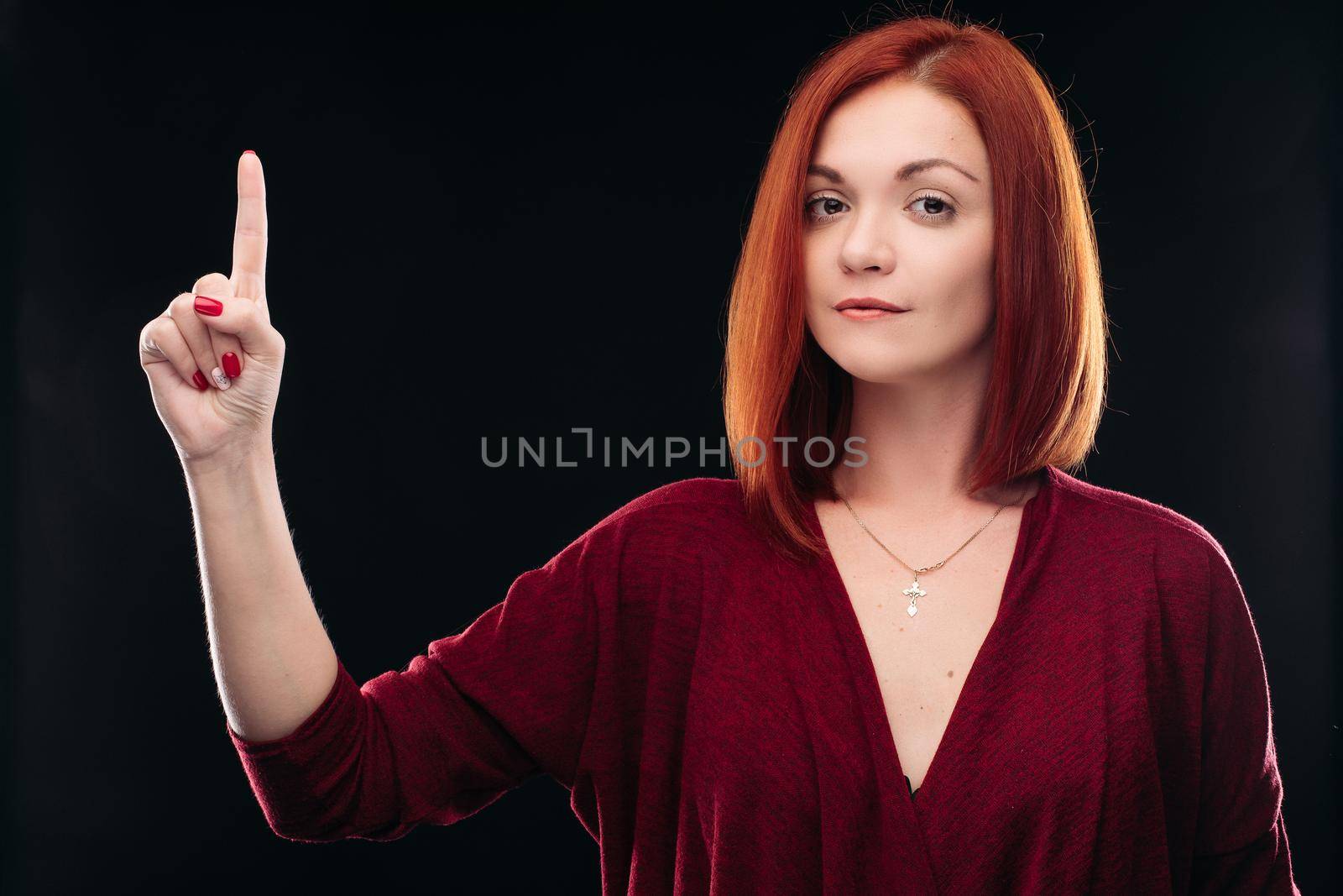 Attractive red haired girl in bordo blouse thinking about something and holding hand with finger up. Young beautiful woman on black background seriously looking at camera. Pretty lady having new idea.