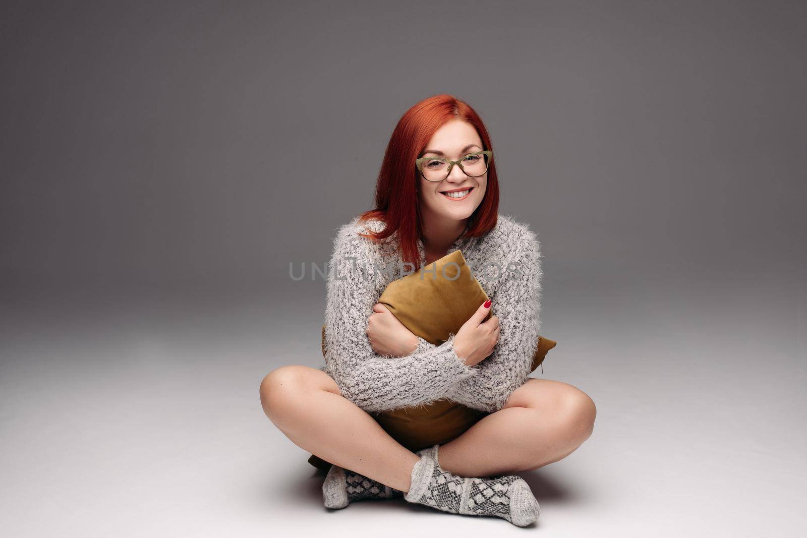 Red haired girl in grey sweater and warm socks sitting on floor in Turkish style and sadly looking at camera. by StudioLucky