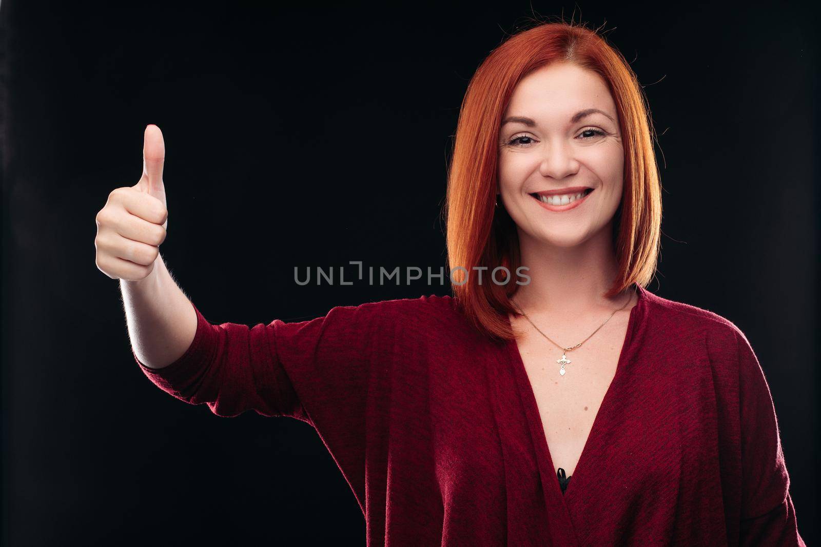 Attractive red haired girl in bordo blouse thinking about something and holding hand with finger up. Young beautiful woman on black background seriously looking at camera. Pretty lady having new idea.