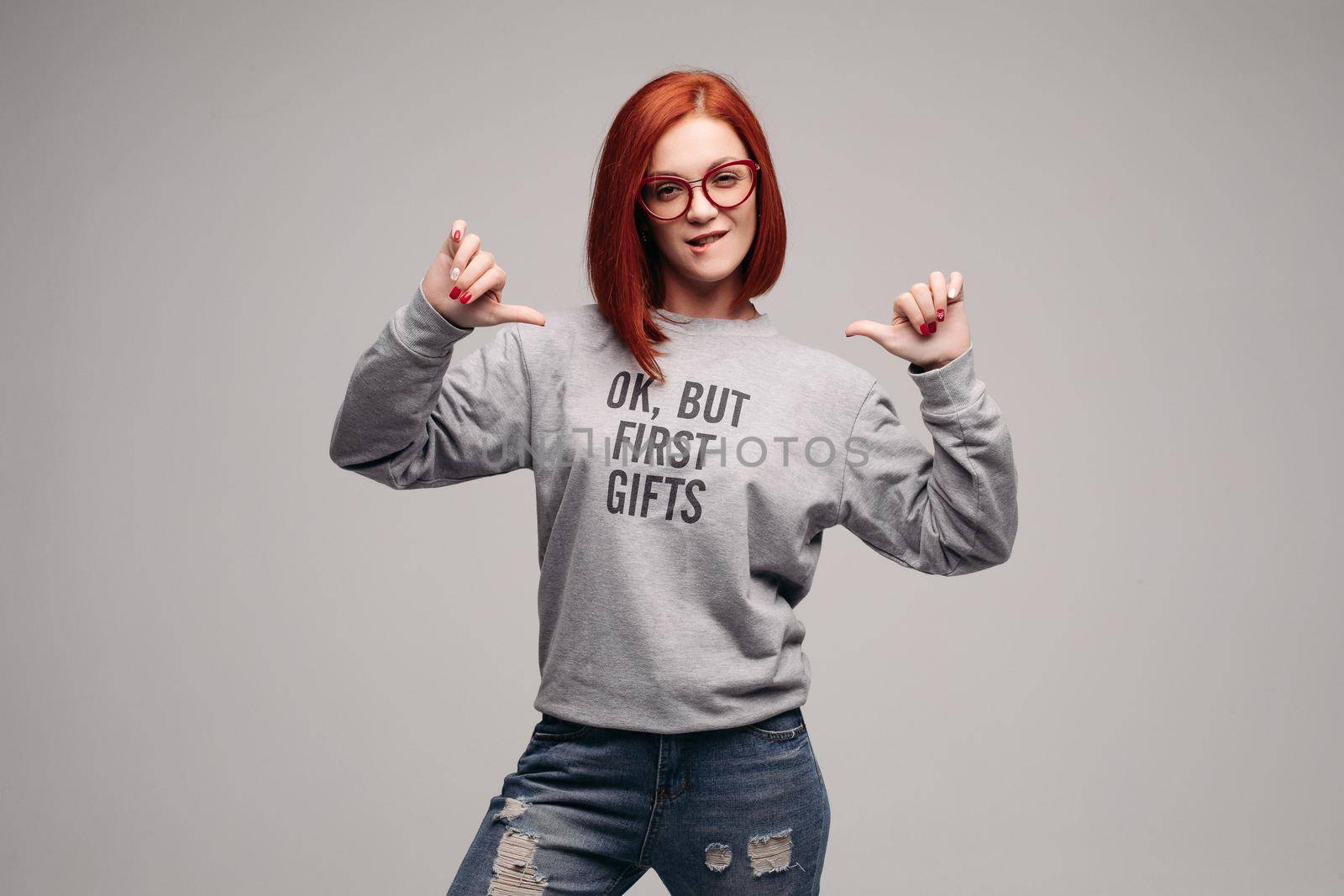 Crop of fashionable red haired girl wearing gray sweatshirt with description about for first gifts. Stylish woman posing in studio pointing by fingers, showing her look, waiting for presents.