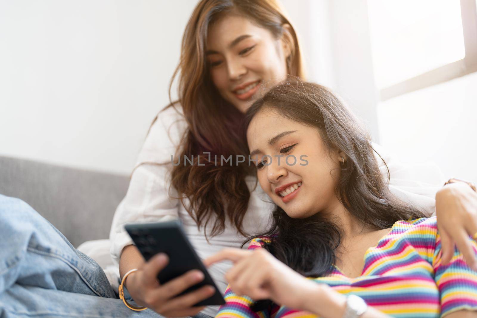 Two happy young aisan casual women having fun using smartphone on couch at home. Lesbian millennial couple. gbt, homosexual, lesbian couple lifestyle