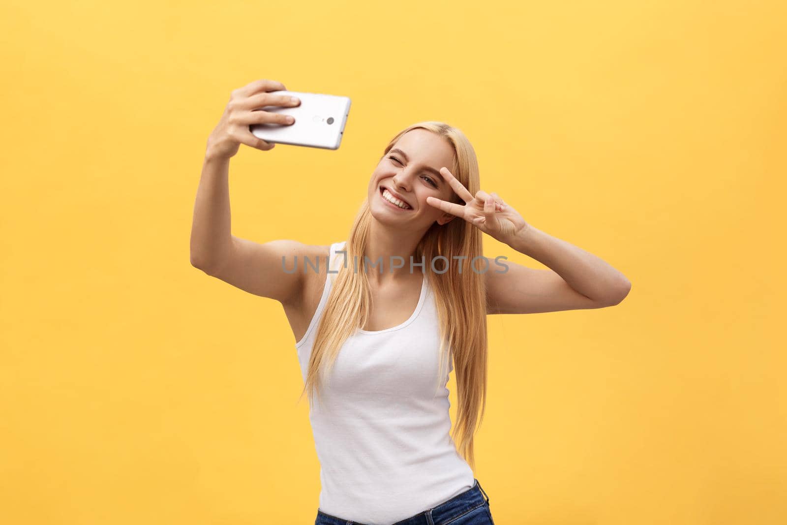 Self portrait of charming cheerful girl shooting selfie on front camera gesturing v-sign peace symbol with fingers isolated on yellow background by Benzoix