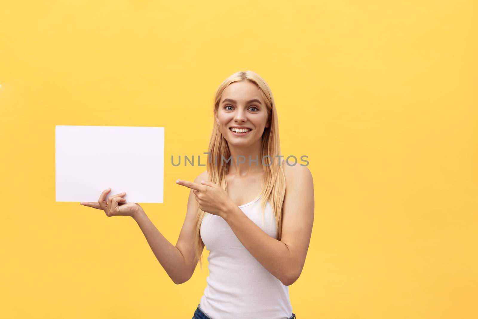 Young woman over yellow background holding blank paper sheet with surprise face pointing finger.