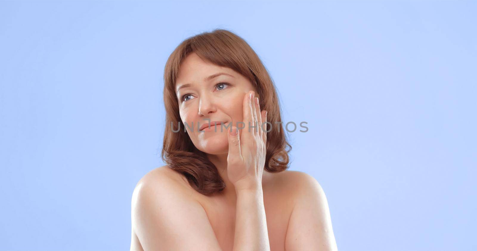 Pretty mature female applies skin care product by touching face with hand. Woman applying anti-age cream on her skin isolated on color background. Spa and wellness concept by LipikStockMedia