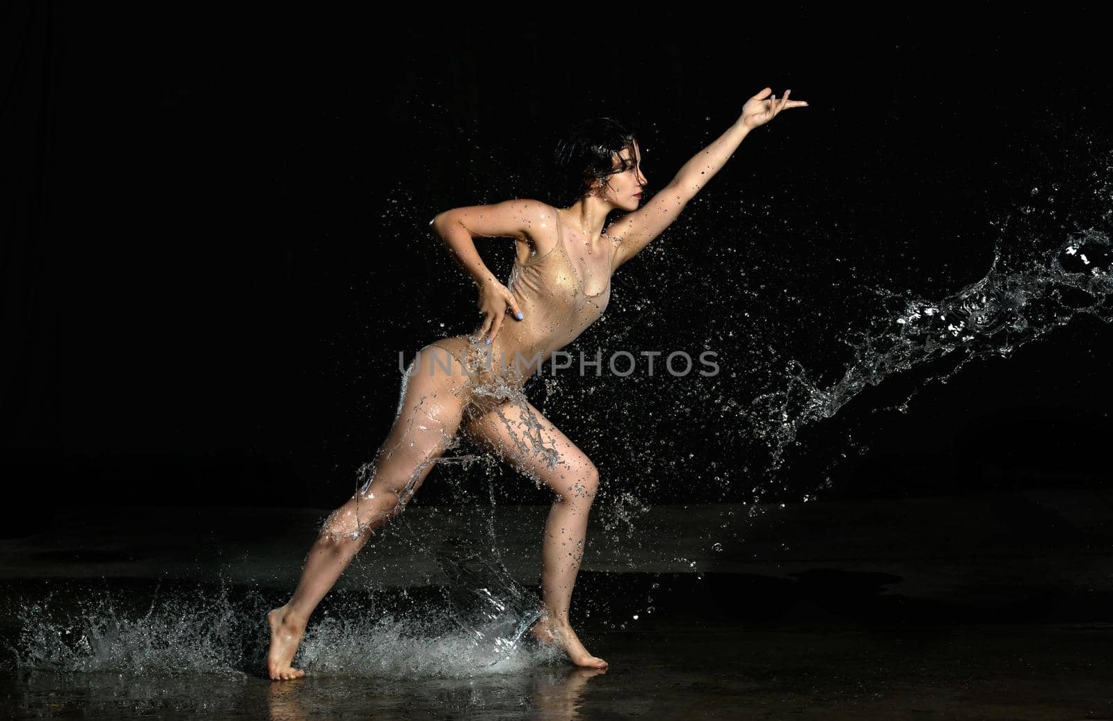 young beautiful woman of Caucasian appearance with long hair dances in drops of water on a black background. Jumping and swinging arms, dressed in a beige bodysuit