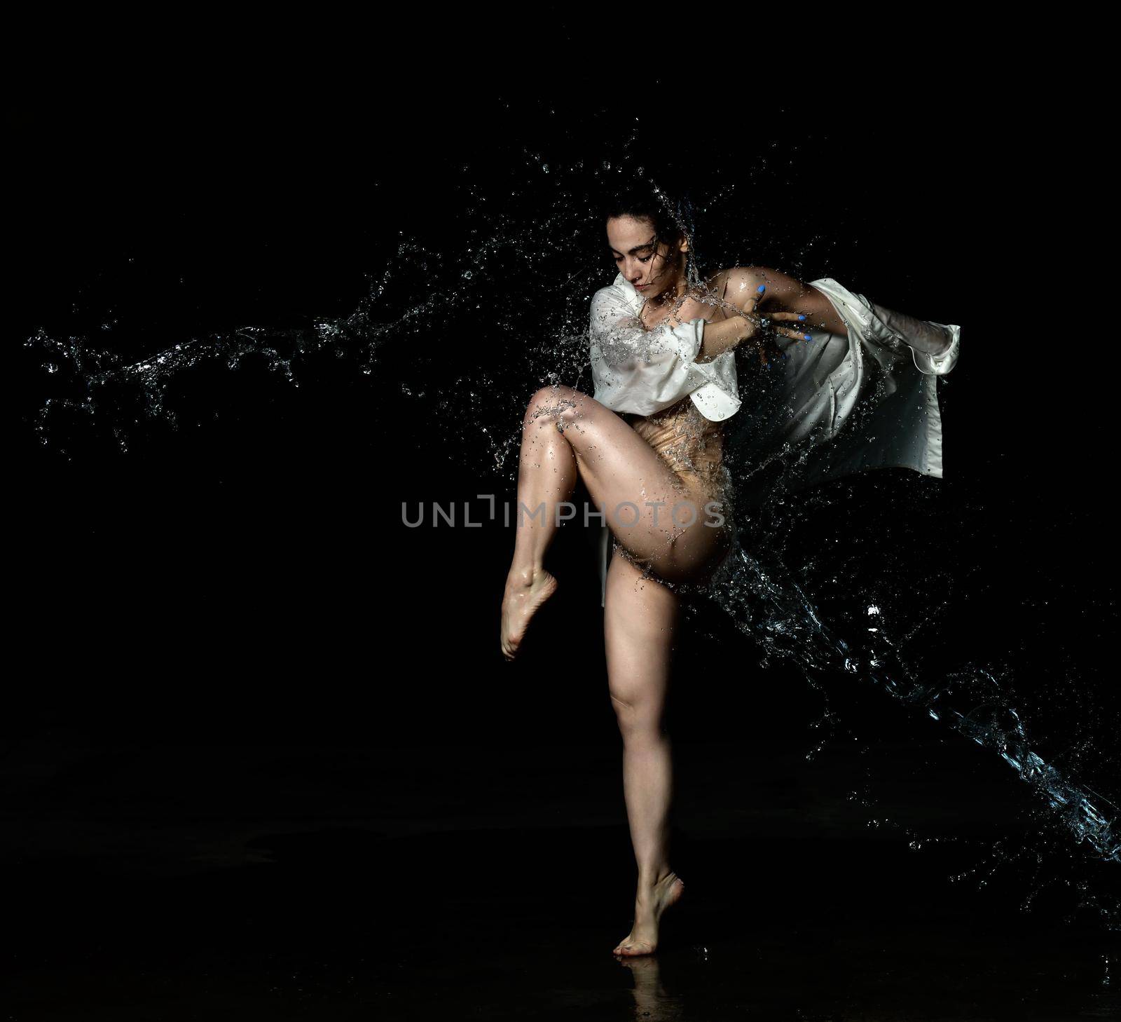 young beautiful woman of Caucasian appearance with long black hair is dancing in a white shirt in the rain on a black background. Sensual and expressive dance, jumping pose