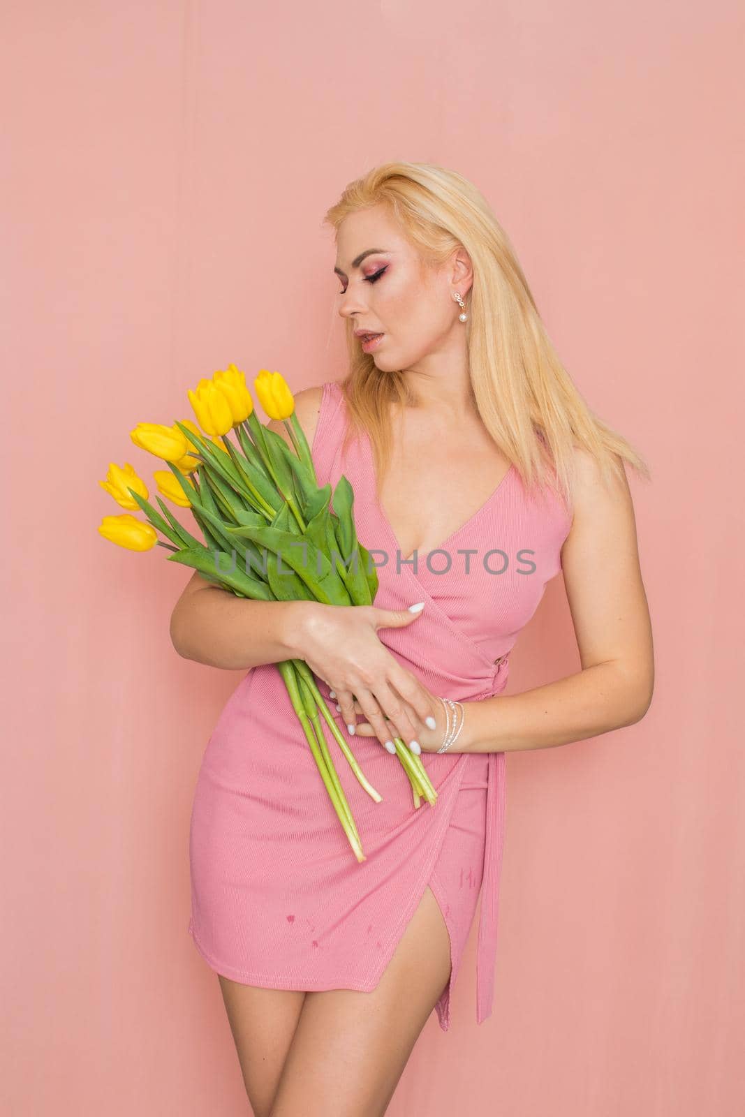 Adult blonde woman in pink dress posing over rosy background. Spring-summer clothing. She is holding bouquet of yellow tulips in her hands. Presents, surprise