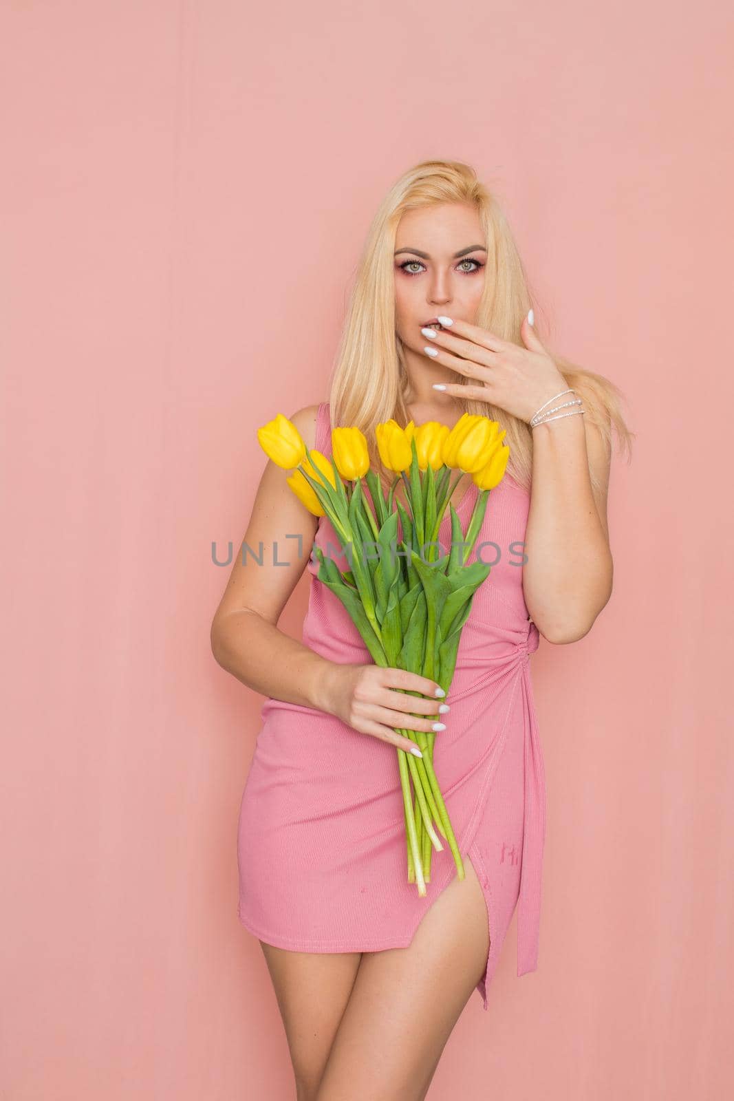 Adult blonde woman in pink dress posing over rosy background. Spring-summer clothing. She is holding bouquet of yellow tulips in her hands. Presents, surprise