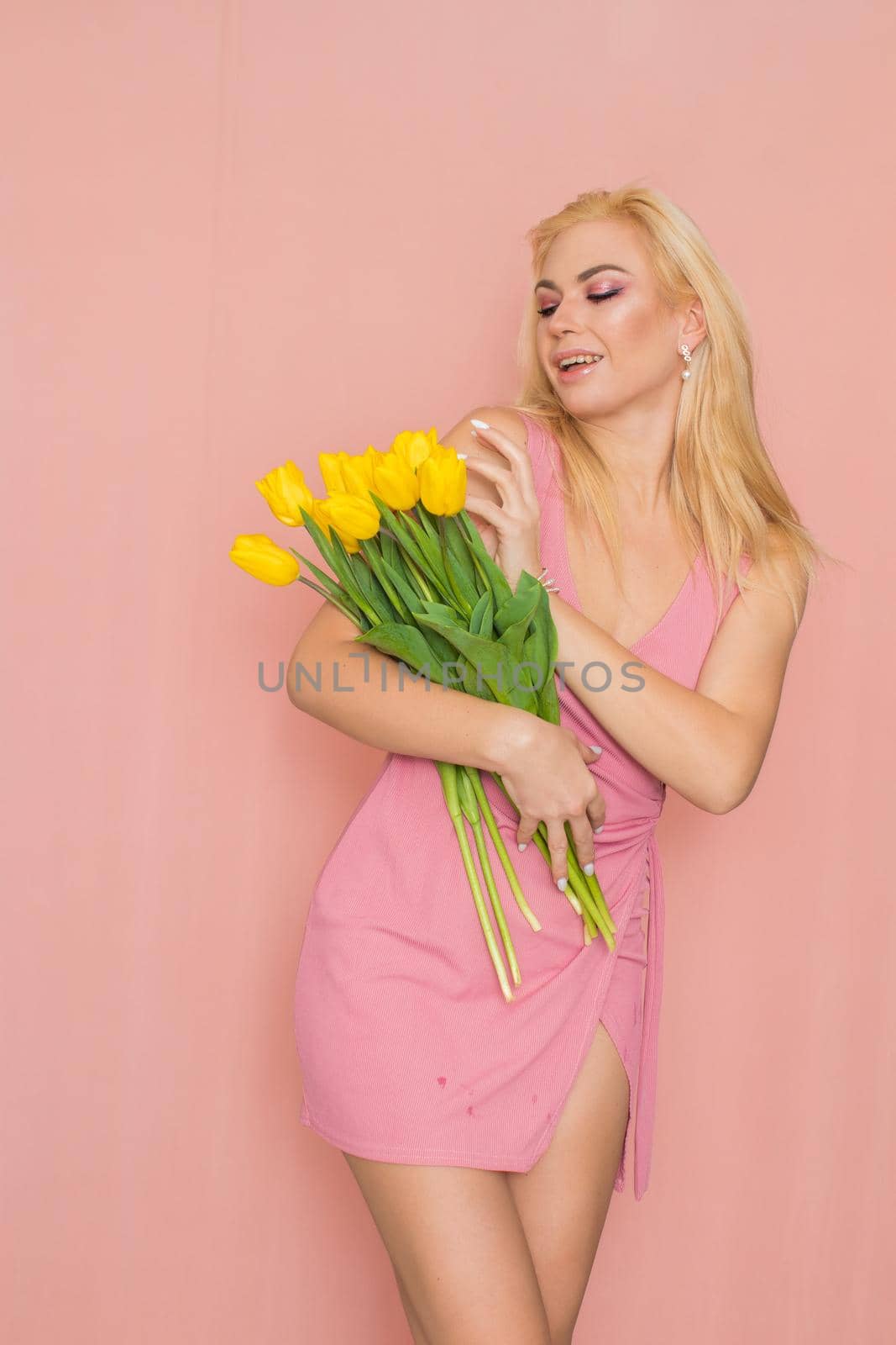 Adult blonde woman in pink dress posing over rosy background. Spring-summer clothing. She is holding bouquet of yellow tulips in her hands. Presents, surprise