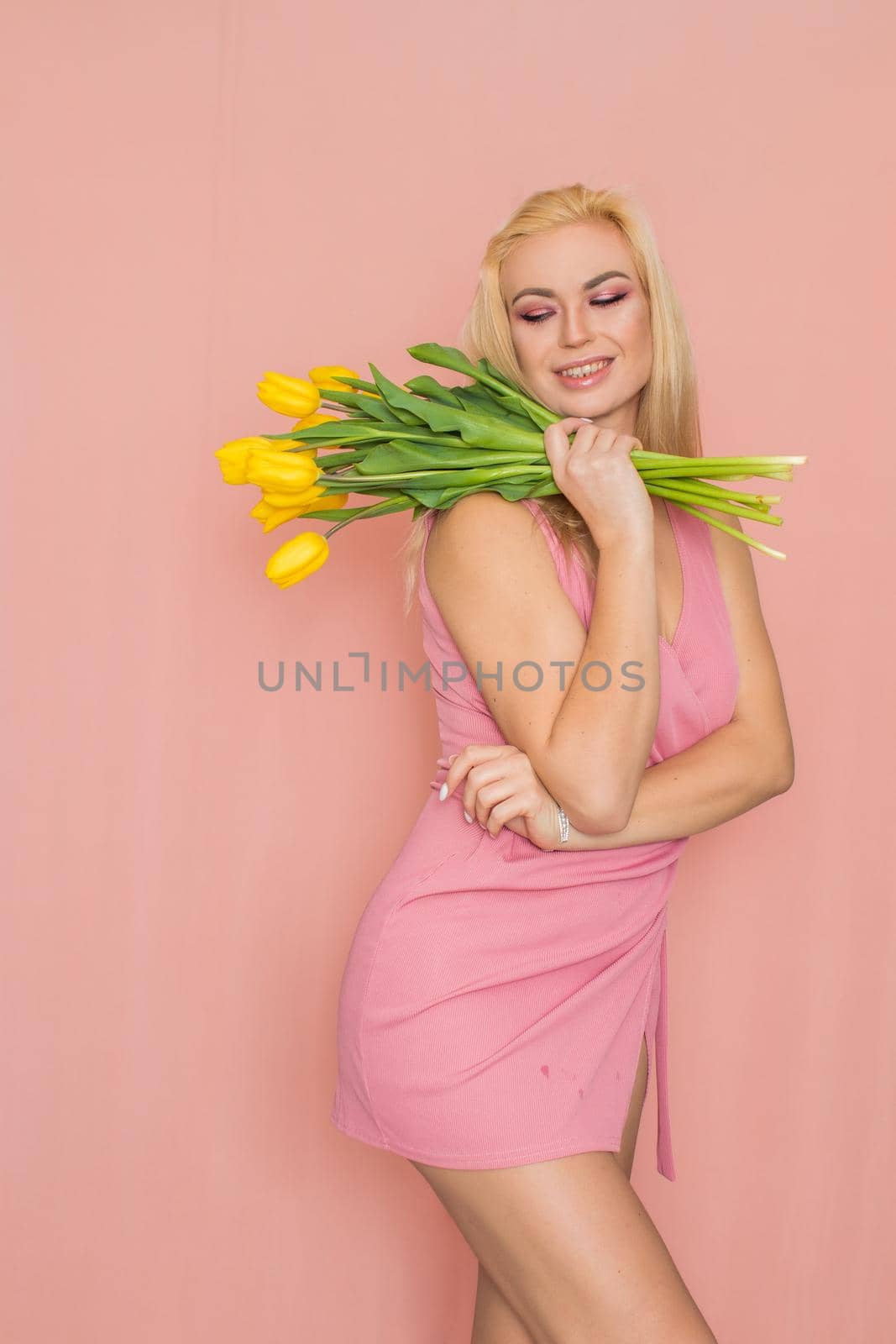 Adult blonde woman in pink dress posing over rosy background. Spring-summer clothing. She is holding bouquet of yellow tulips in her hands. Presents, surprise