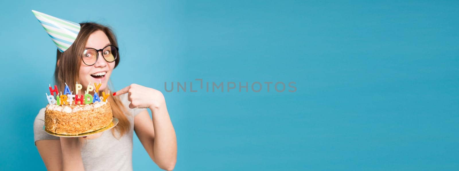 Charming merry crazy young girl student in congratulatory paper hat holding a happy birthday cake in her hands standing against a blue background. Advertising space