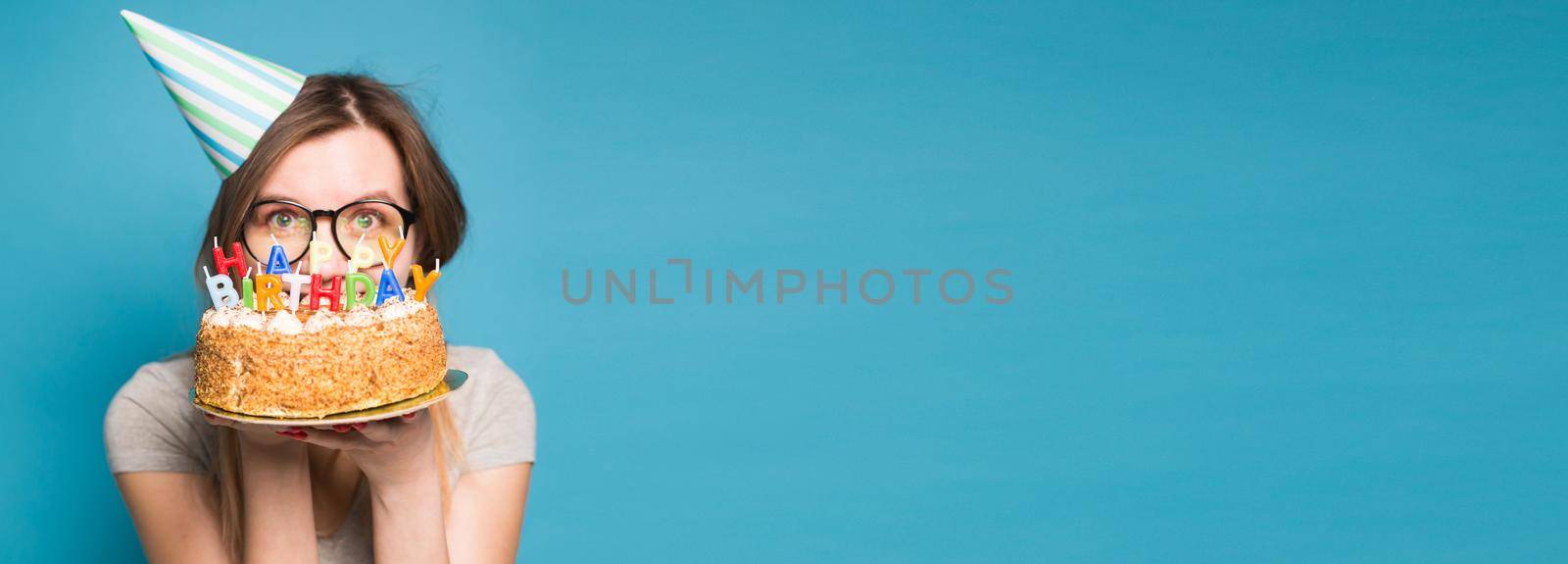 Banner with funny positive girl in glasses and greeting paper hat holding a happy birthday cake in her hands standing on blue background with copy space and empty space by Satura86