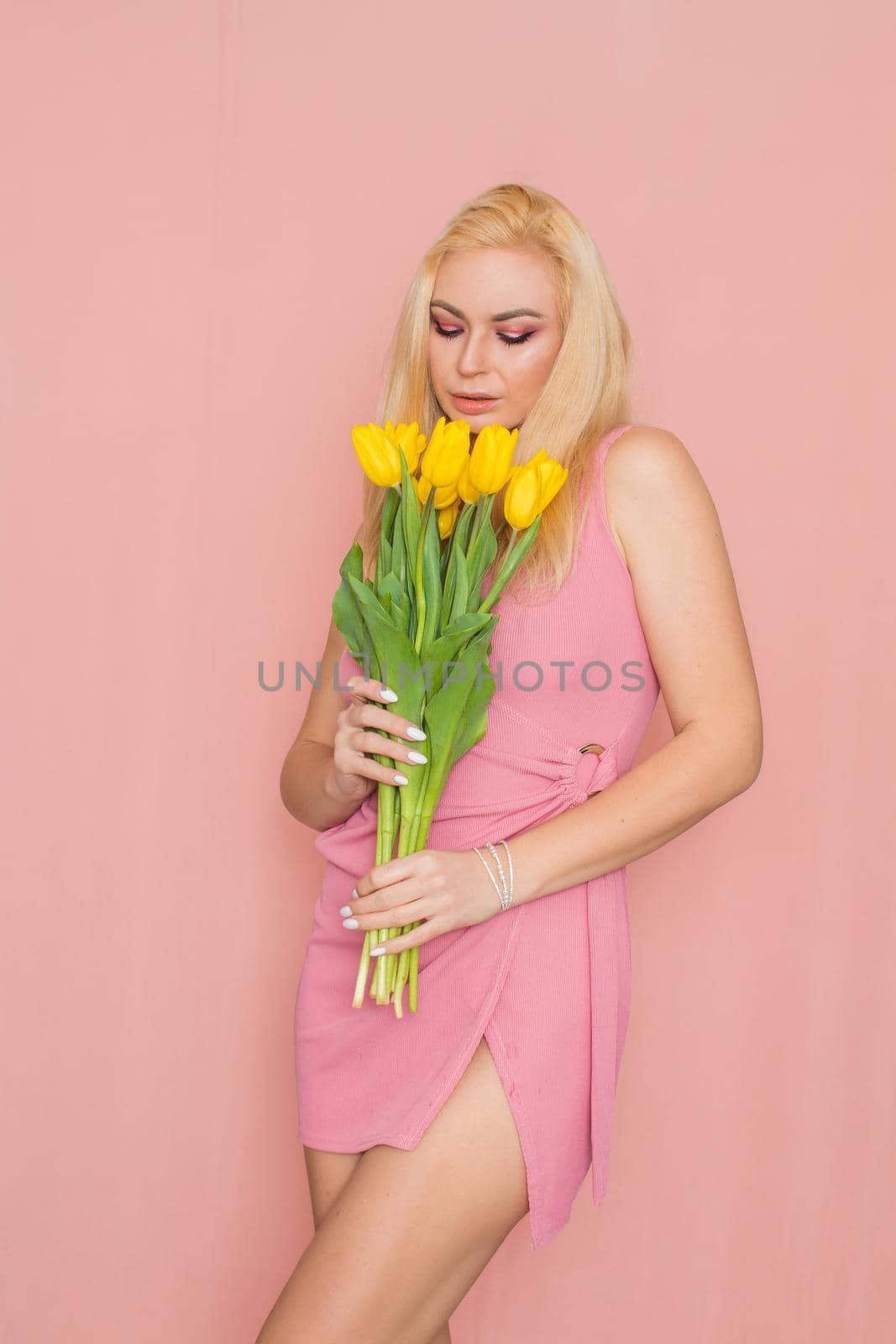 Adult blonde woman in pink dress posing over rosy background. Spring-summer clothing. She is holding bouquet of yellow tulips in her hands. Presents, surprise