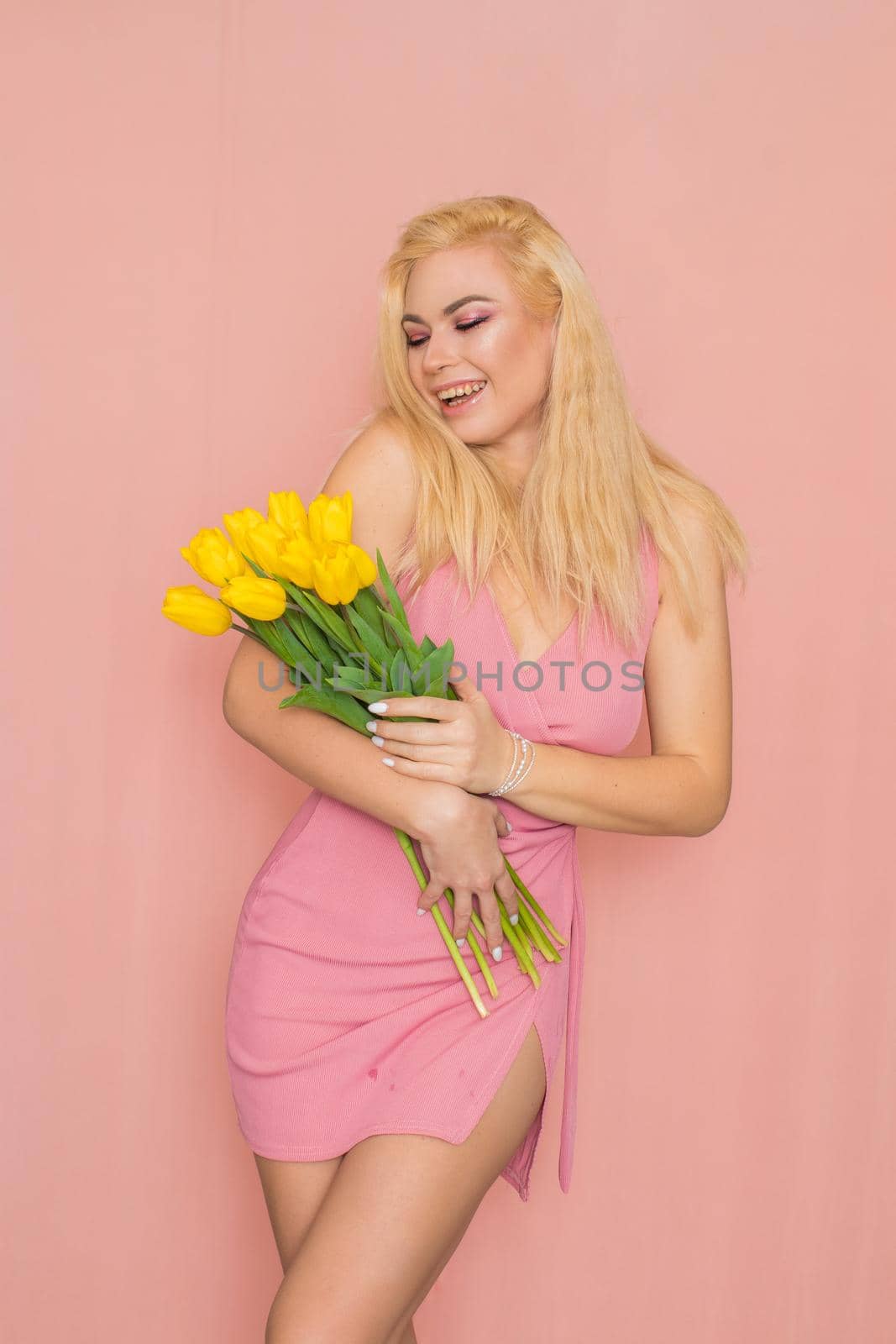 Adult blonde woman in pink dress posing over rosy background. Spring-summer clothing. She is holding bouquet of yellow tulips in her hands. Presents, surprise