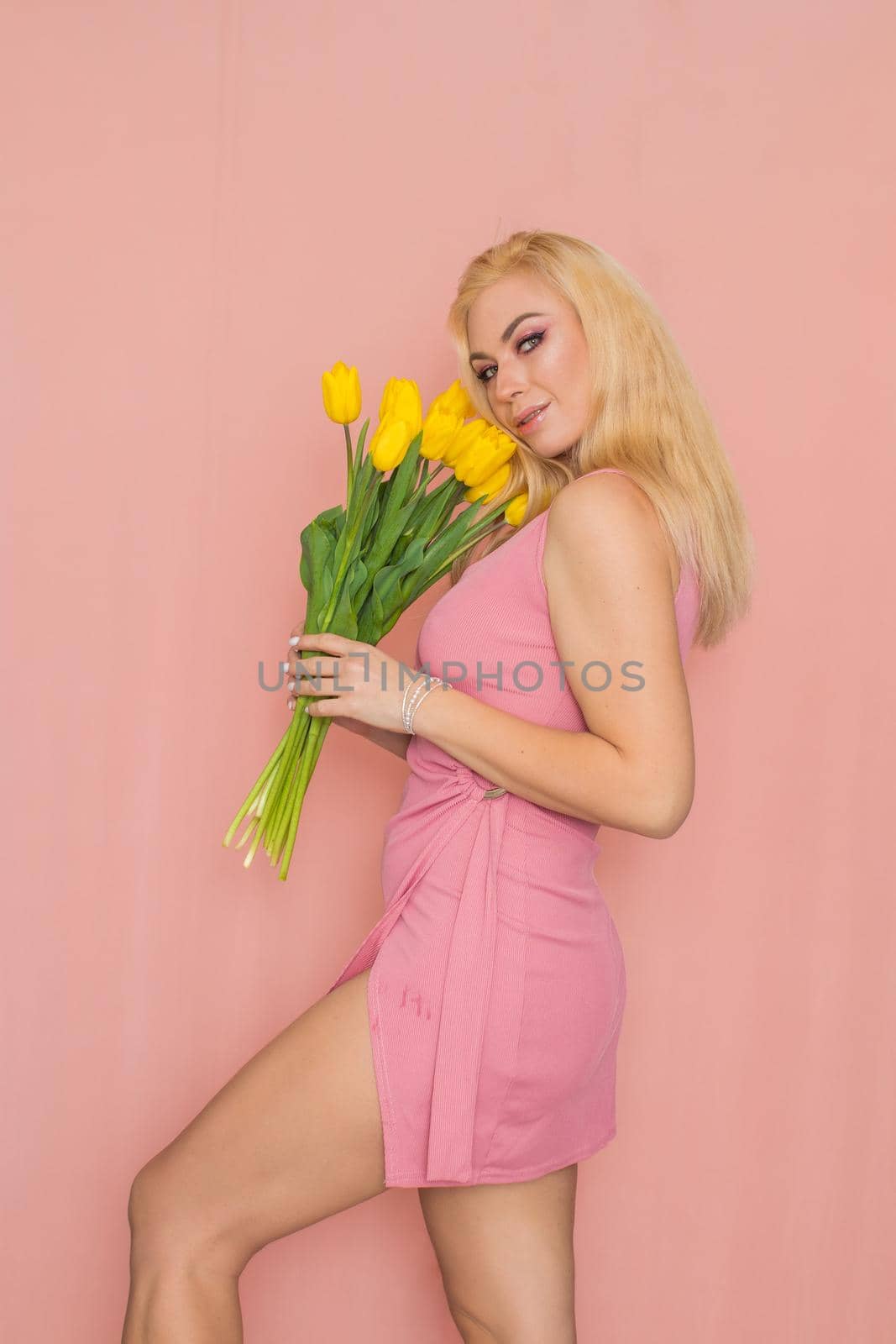 Adult blonde woman in pink dress posing over rosy background. Spring-summer clothing. She is holding bouquet of yellow tulips in her hands. Presents, surprise