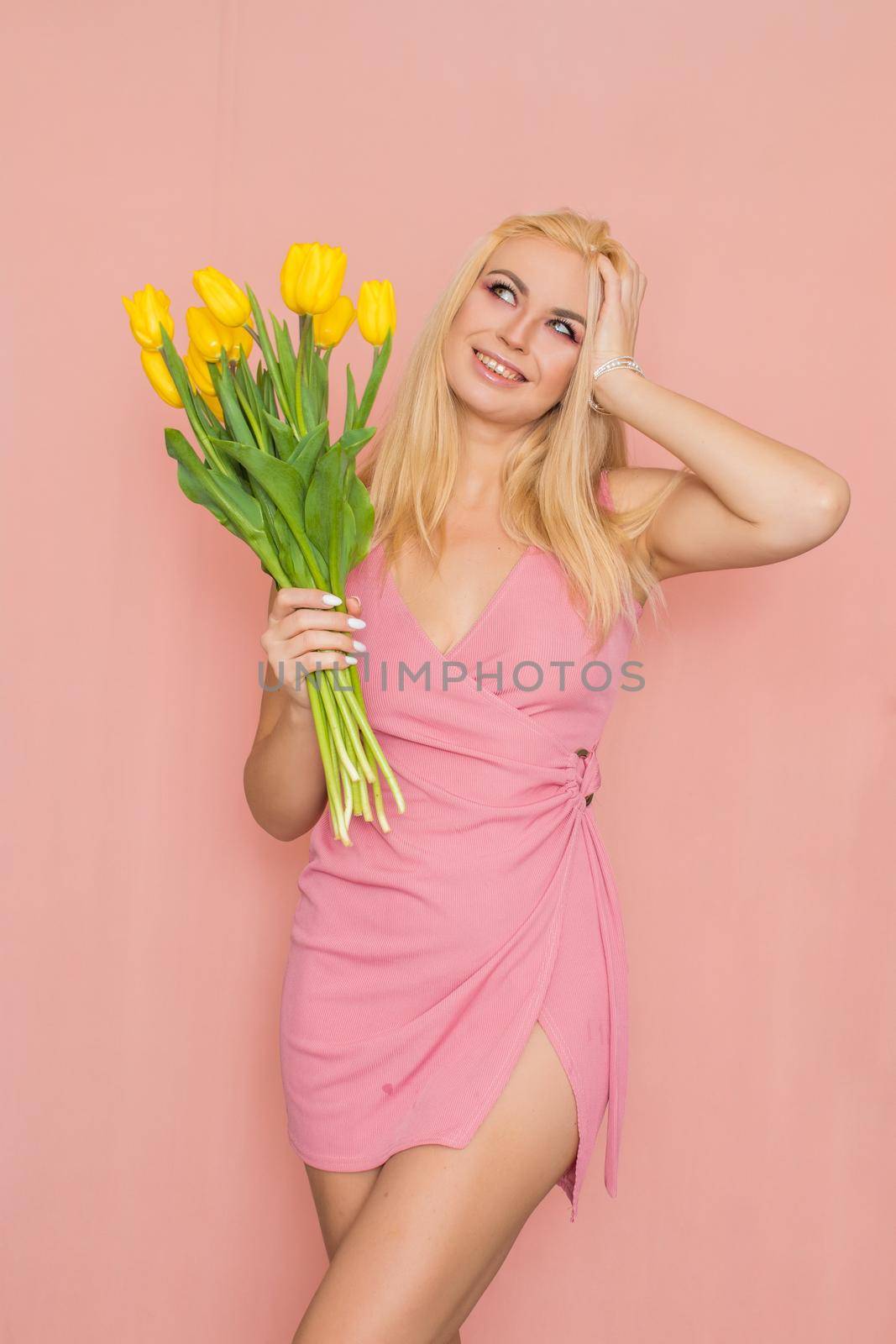 Adult blonde woman in pink dress posing over rosy background. Spring-summer clothing. She is holding bouquet of yellow tulips in her hands. Presents, surprise