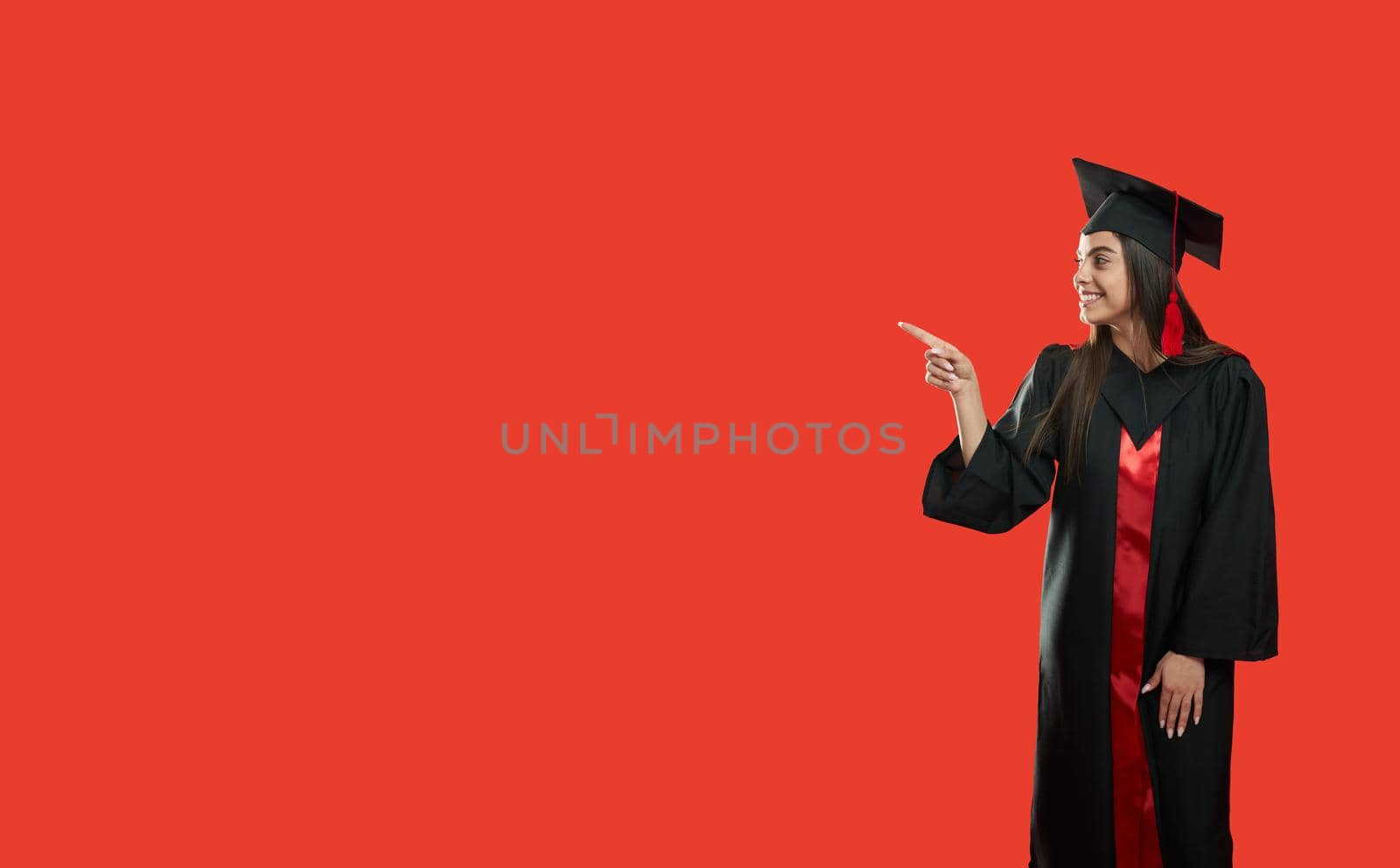 Sise view of beautiful girl in graduate gown and mortarboard standing, showing by finger. Young slim female grauduating from college, university. Isolatedon red background.
