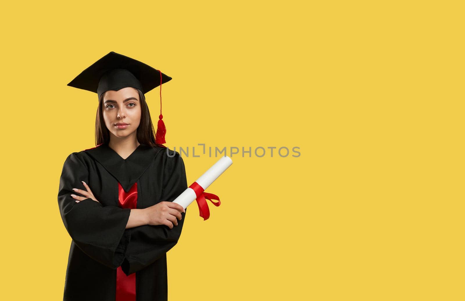 Front view of girl standing with crossed hands, holding diploma. Brunette graduating from college, wearing mortarboard and graduate gown, looking at camera. Concept of youth.