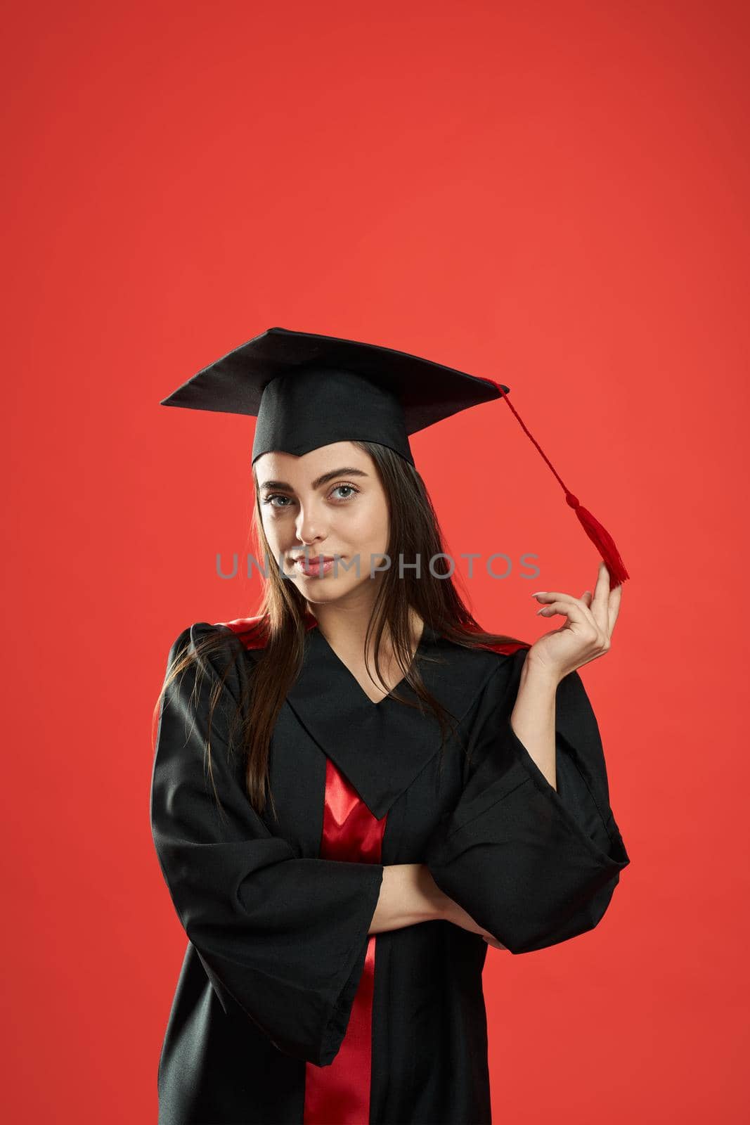 Front view of pretty girl in mortarboard and graduate gown standing with crossed hands. Pretty female student smiling, looking at camera. Isolated on red studio background,