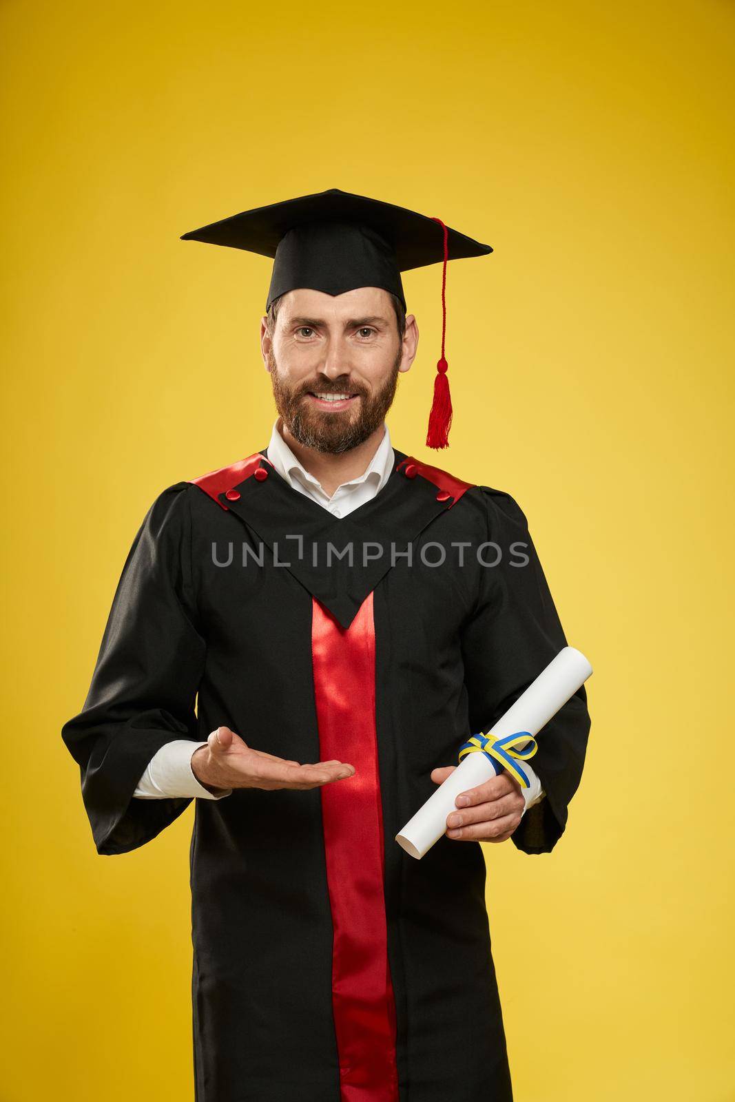 Front view of proud student showing diploma. Handsome man wearing graduate gown and mortarboard, looking at camera, cheerful, happy. Isolated on yellow studio background.