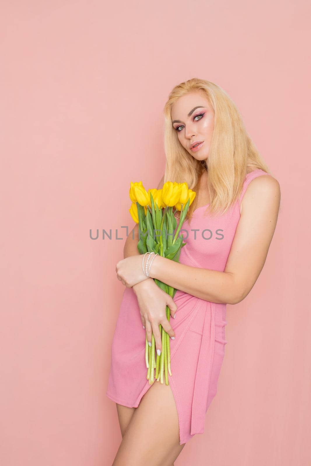 Adult blonde woman in pink dress posing over rosy background. Spring-summer clothing. She is holding bouquet of yellow tulips in her hands. Presents, surprise