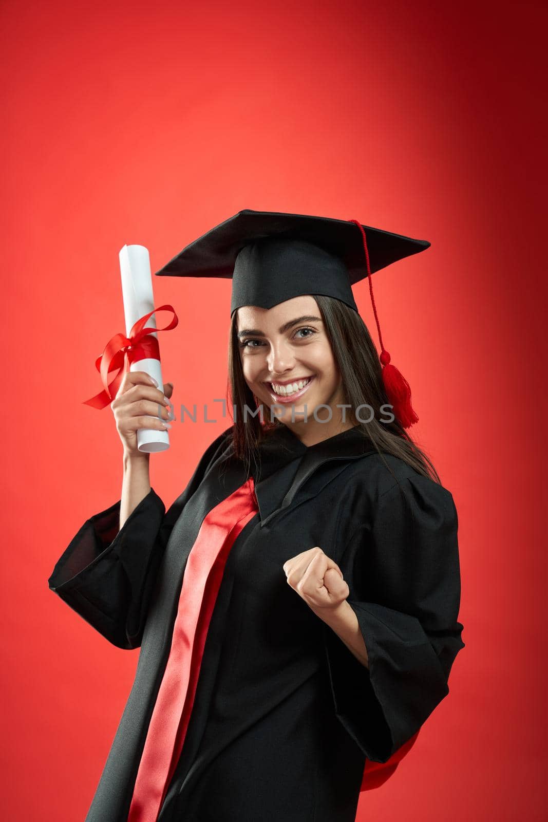 Front view of pretty young female holding diploma. Girl smiling, looking at camera, showing hurray, wearing graduate gown and mortarboard. Concept of education and youth.