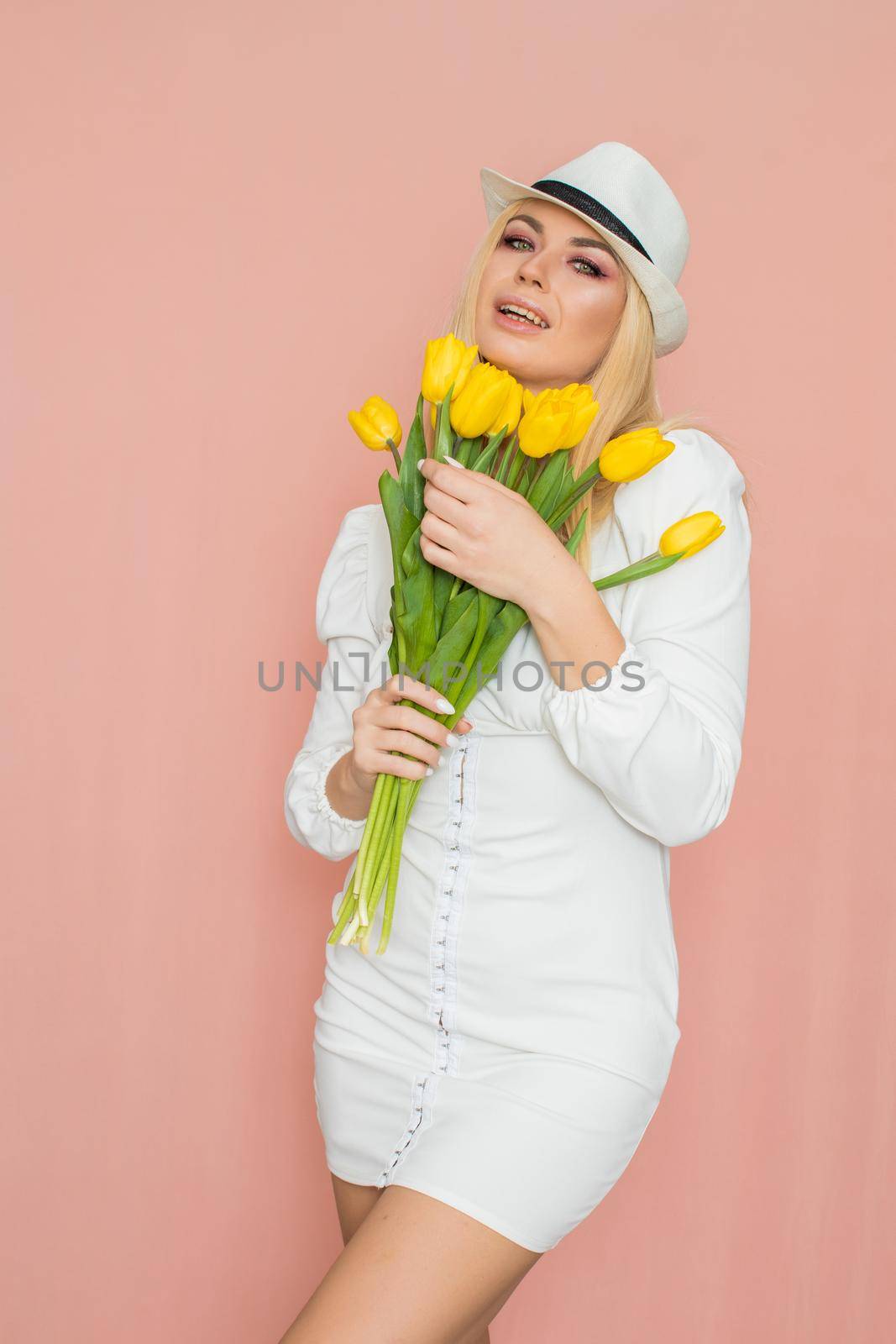 Spring fashion photo of blonde woman posing over pink background. Wearing white vintage dress with long sleeves and white hat. Holding bouquet with yellow tulips in her hands