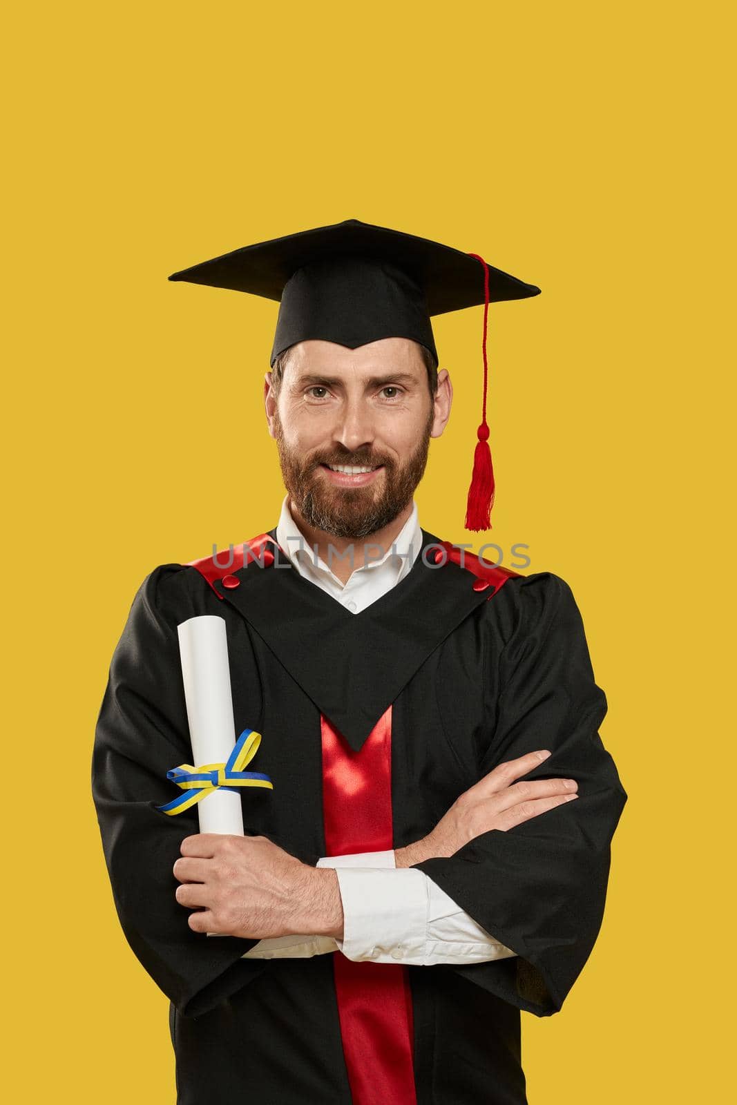 Front view of man with beard graduating from university, college. Graduate wearing mortarboard standing with crossed hands, smiling, holding diploma. Concept of high school.