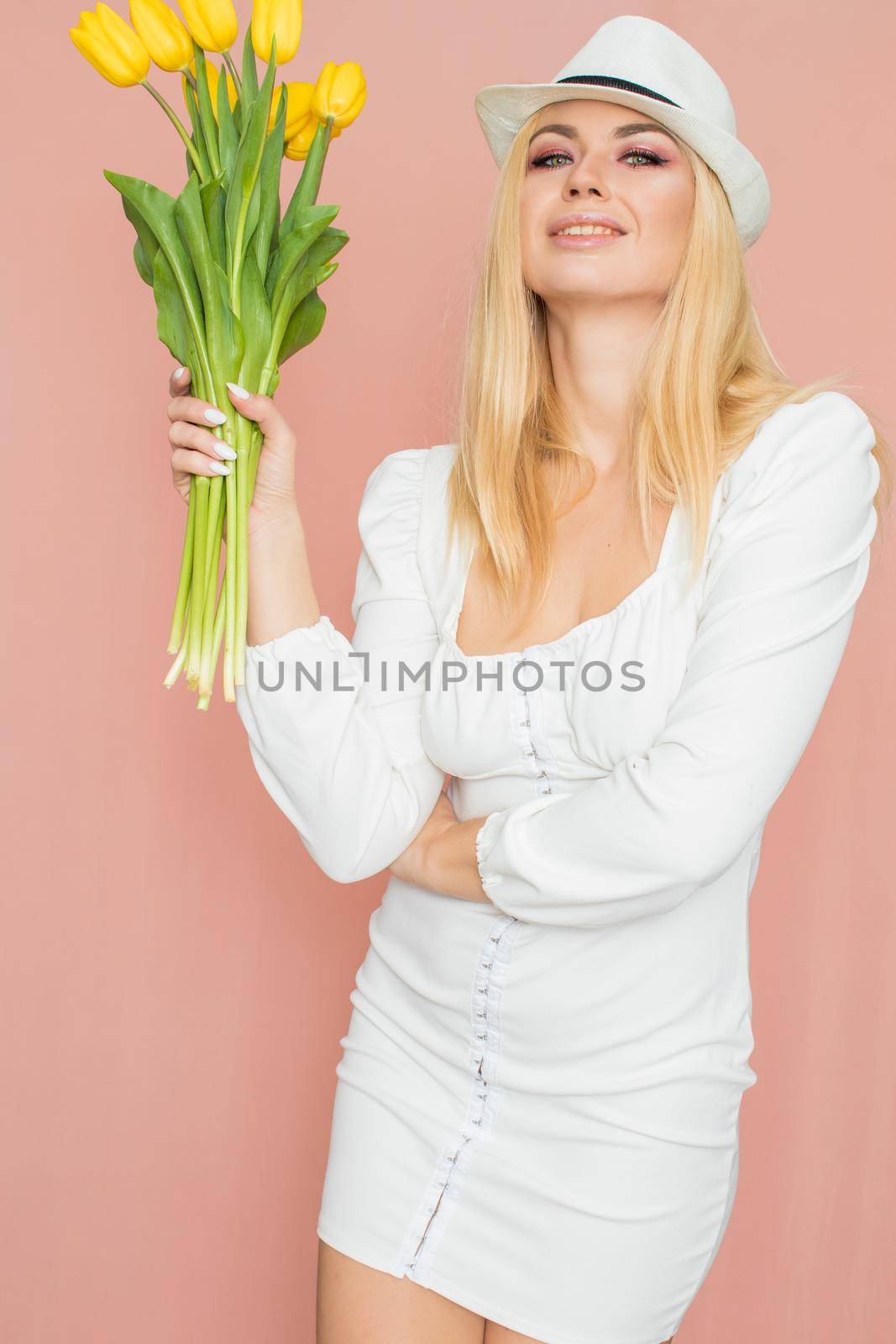 Spring fashion photo of blonde woman posing over pink background. Wearing white vintage dress with long sleeves and white hat. Holding bouquet with yellow tulips in her hands