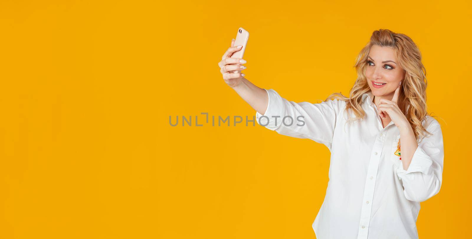 Portrait of a stylish beautiful smiling caucasian blonde woman taking a selfie on a smartphone, holding a mobile phone in outstretched hand and trying to look beautiful in the photo, yellow background