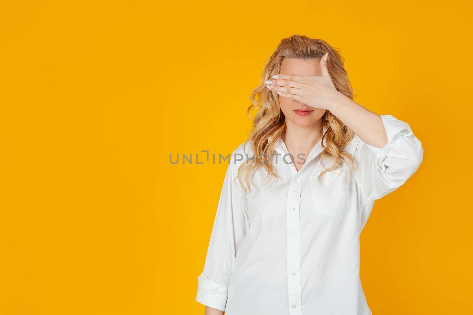 Portrait of a woman closes her eyes with the palm of her hand. Blonde waiting for an unexpected gift, waiting for the present, counting ten, playing hide and seek, standing on a yellow background
