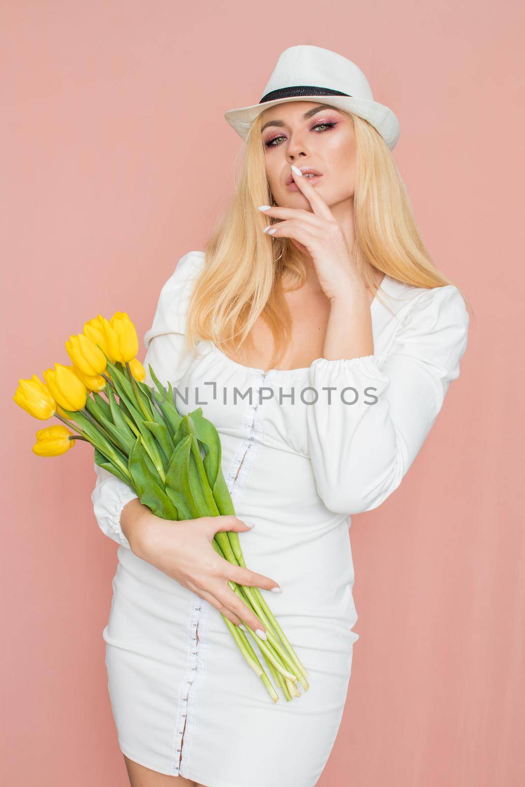 Spring fashion photo of blonde woman posing over pink background. Wearing white vintage dress with long sleeves and white hat. Holding bouquet with yellow tulips in her hands