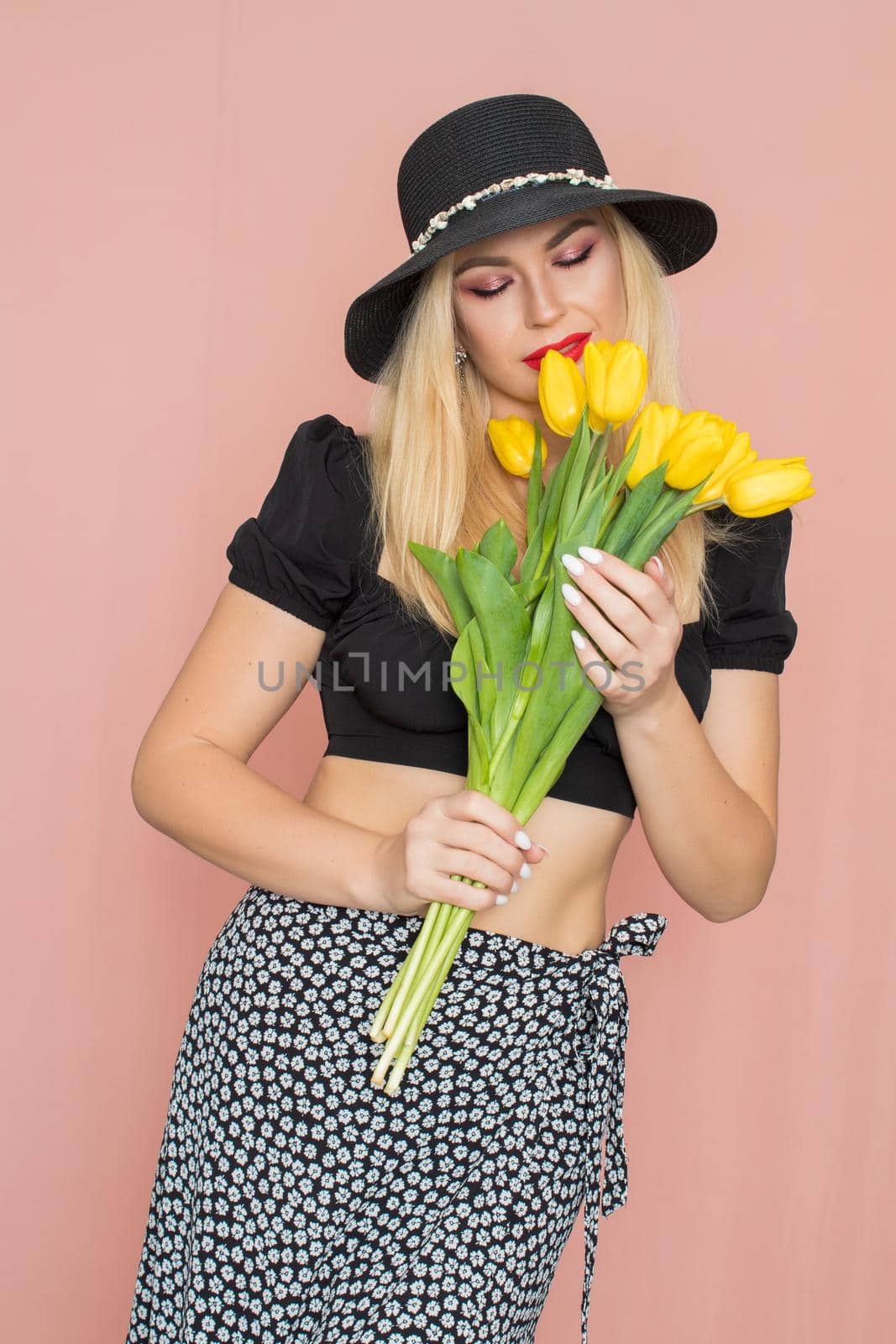 Summer fashion portrait blonde woman. Sexy look in black top and skirt, wearing hat. Red lips. Holding yellow tulips in her hands