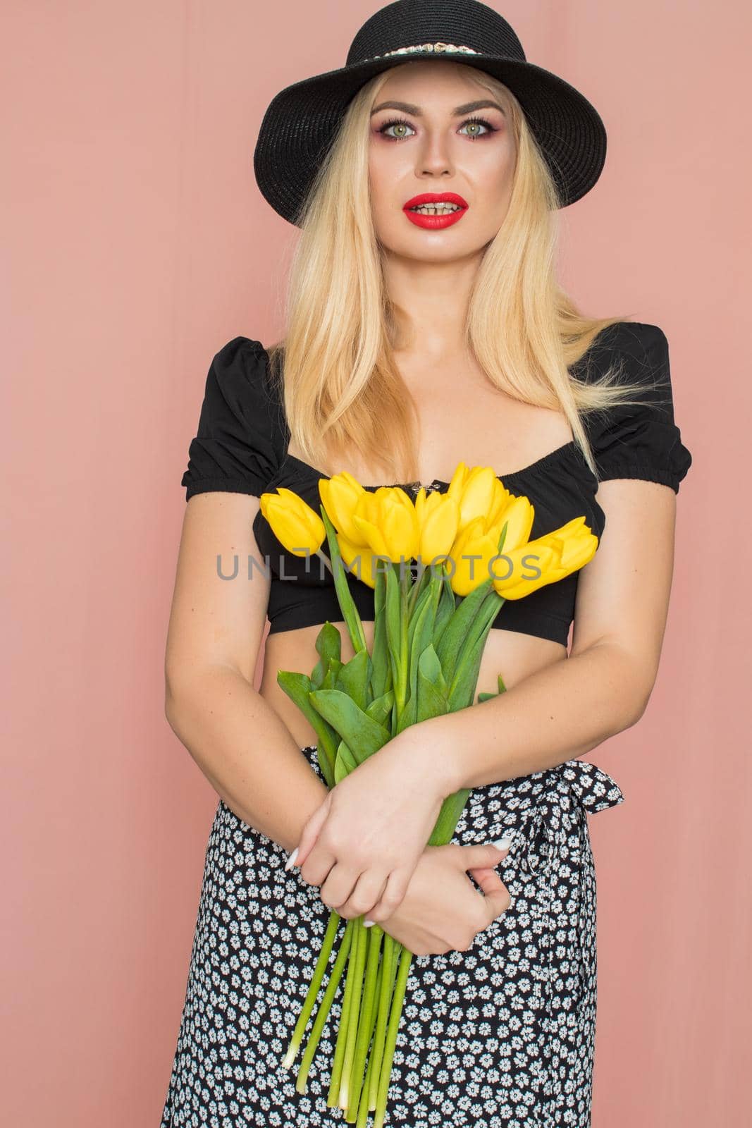 Summer fashion portrait blonde woman. Sexy look in black top and skirt, wearing hat. Red lips. Holding yellow tulips in her hands