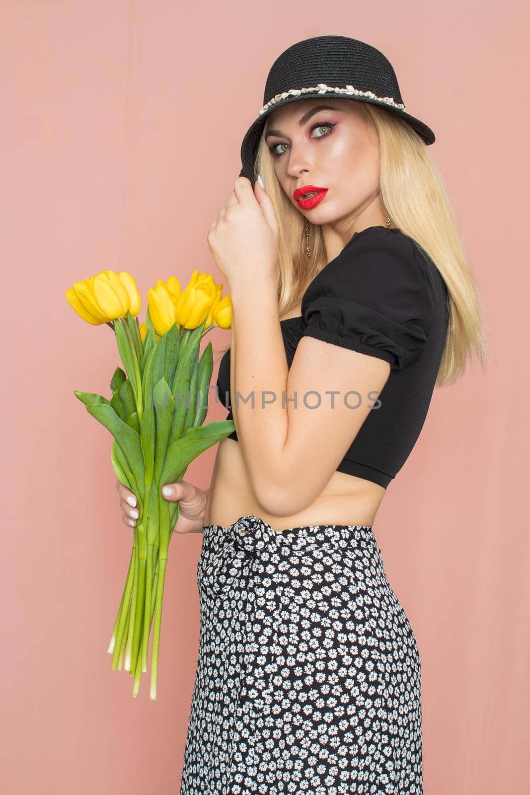 Summer fashion portrait blonde woman. Sexy look in black top and skirt, wearing hat. Red lips. Holding yellow tulips in her hands
