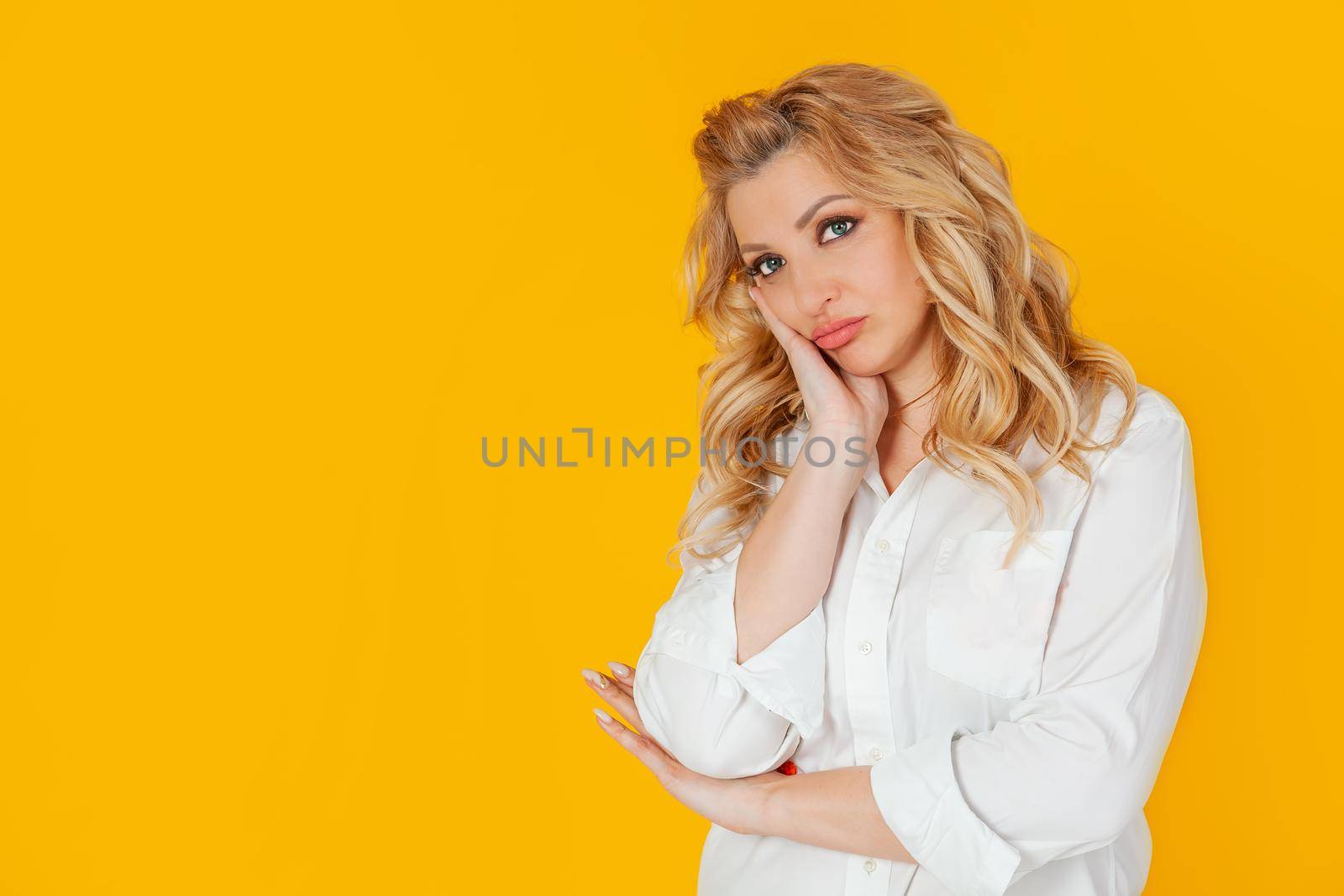 The blonde woman is bored, feels sleepy and reluctantly looks at the camera, holds a palm on her cheek disinterestedly listening to a boring story with a bored face, standing on a yellow background