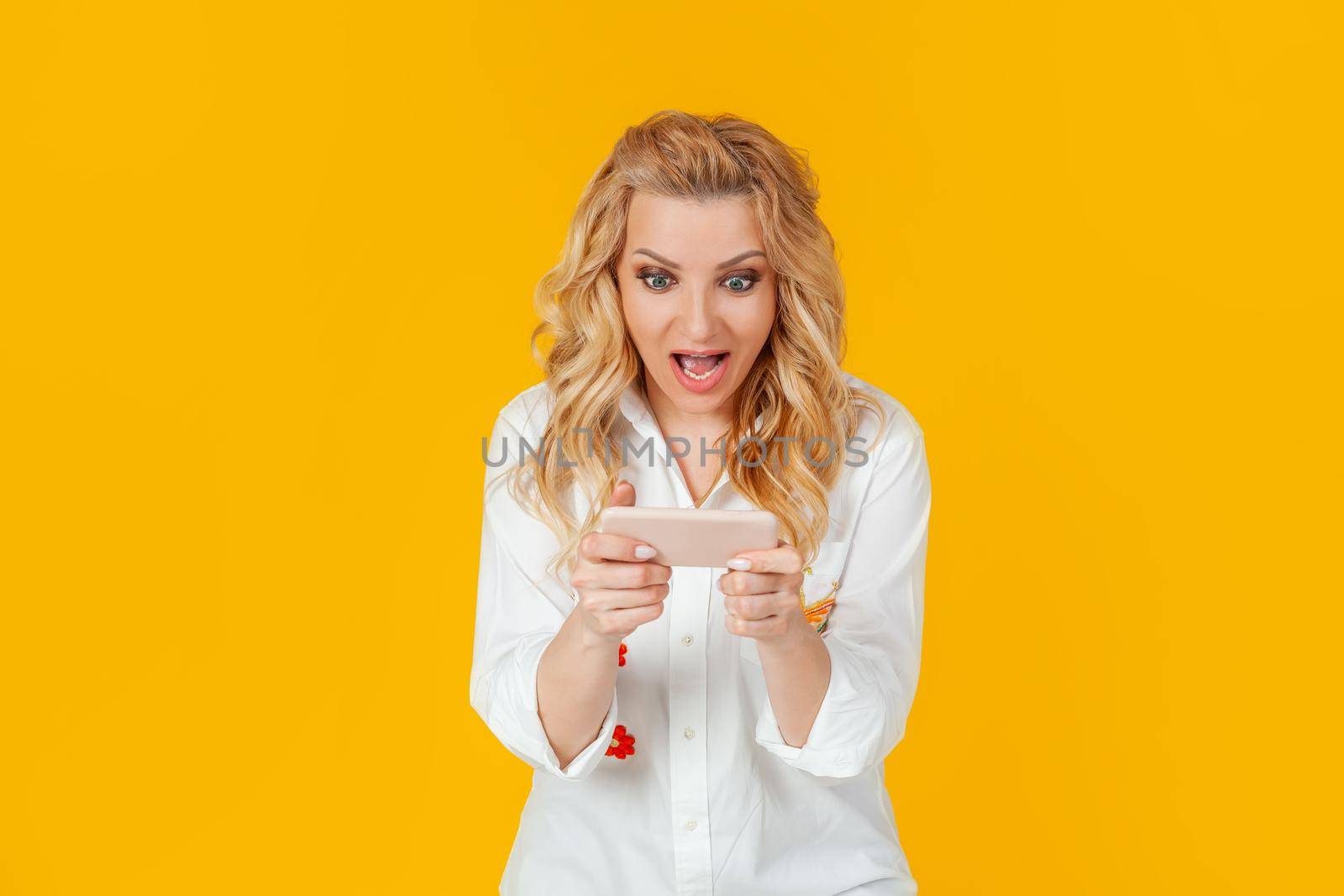 Cheerful excited pretty woman plays a new amazing game for smartphones, screams joyfully and smiles, winning, on a yellow background