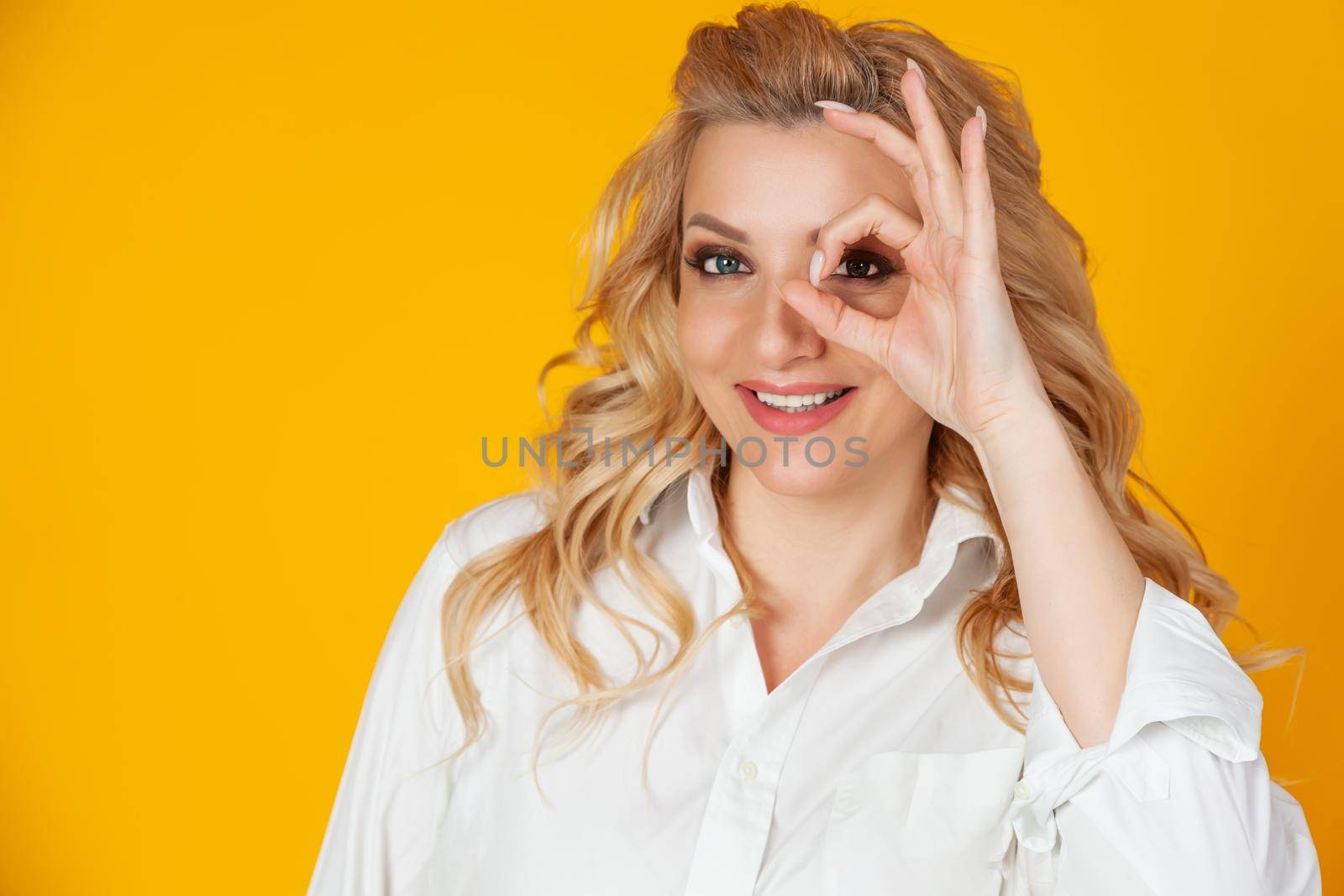 Blond woman feeling optimistic happy showing ok circle over eye smiling happily. feels happy optimistic showing ok, good gesture okay with an eye sign smiling happily standing yellow background