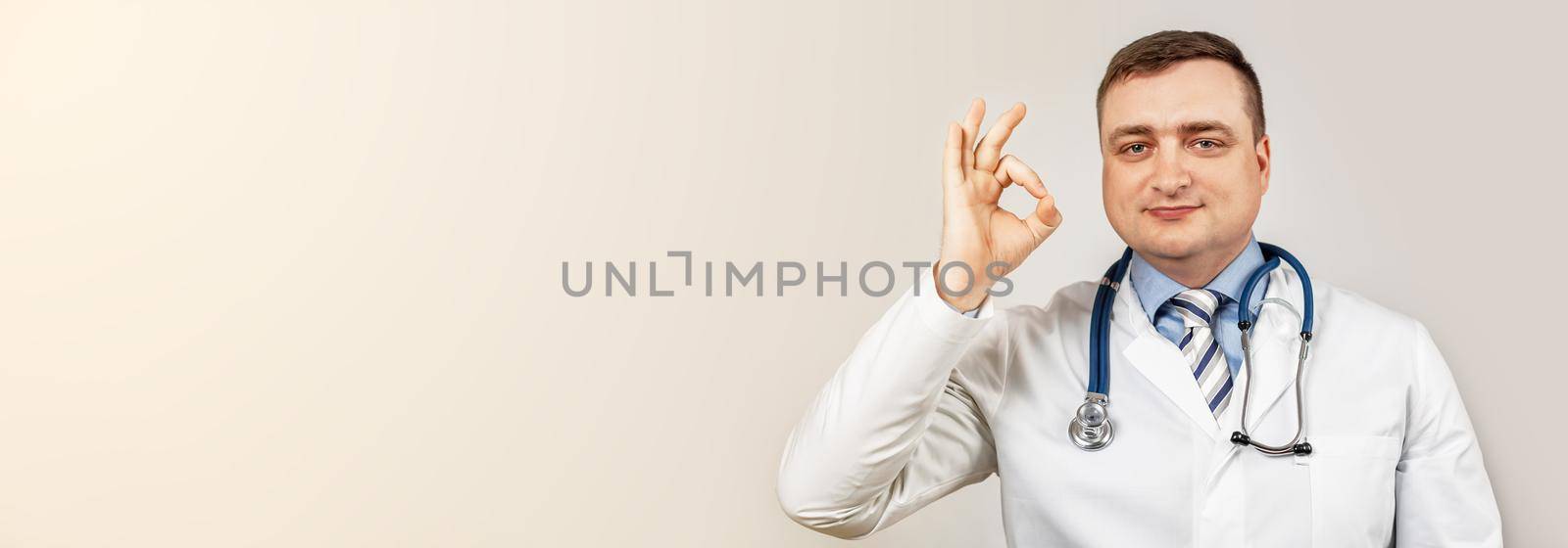 Good job. Satisfied doctor man in a medical coat and with a stethoscope shows okay okay sign of approval smiles, white background. No problem, clinic guarantee best professional service and caring.