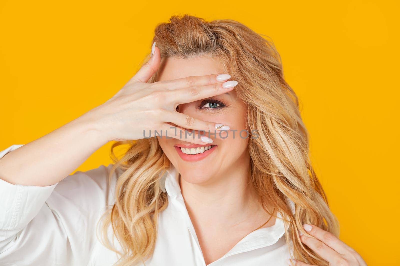 Portrait of a beautiful enthusiastic woman with curly blond hair closes her eyes and looks through her fingers, smiling. Looking to the future with an optimistic look.