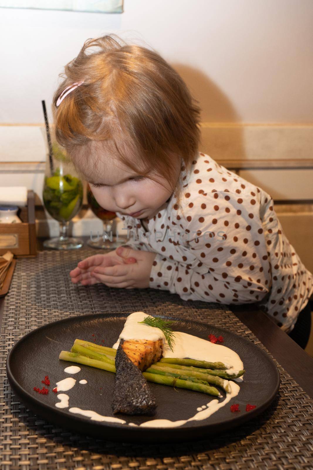 A little girl in a restaurant looks at what the official brought on a plate with baked salmon and asparagus, decorated with sauce and caviar of flying fish, on the table in front of her she looks very carefully at how the fish dish is decorated Her daughter looks at the food with hungry eyes and wants to try it as quickly as possible