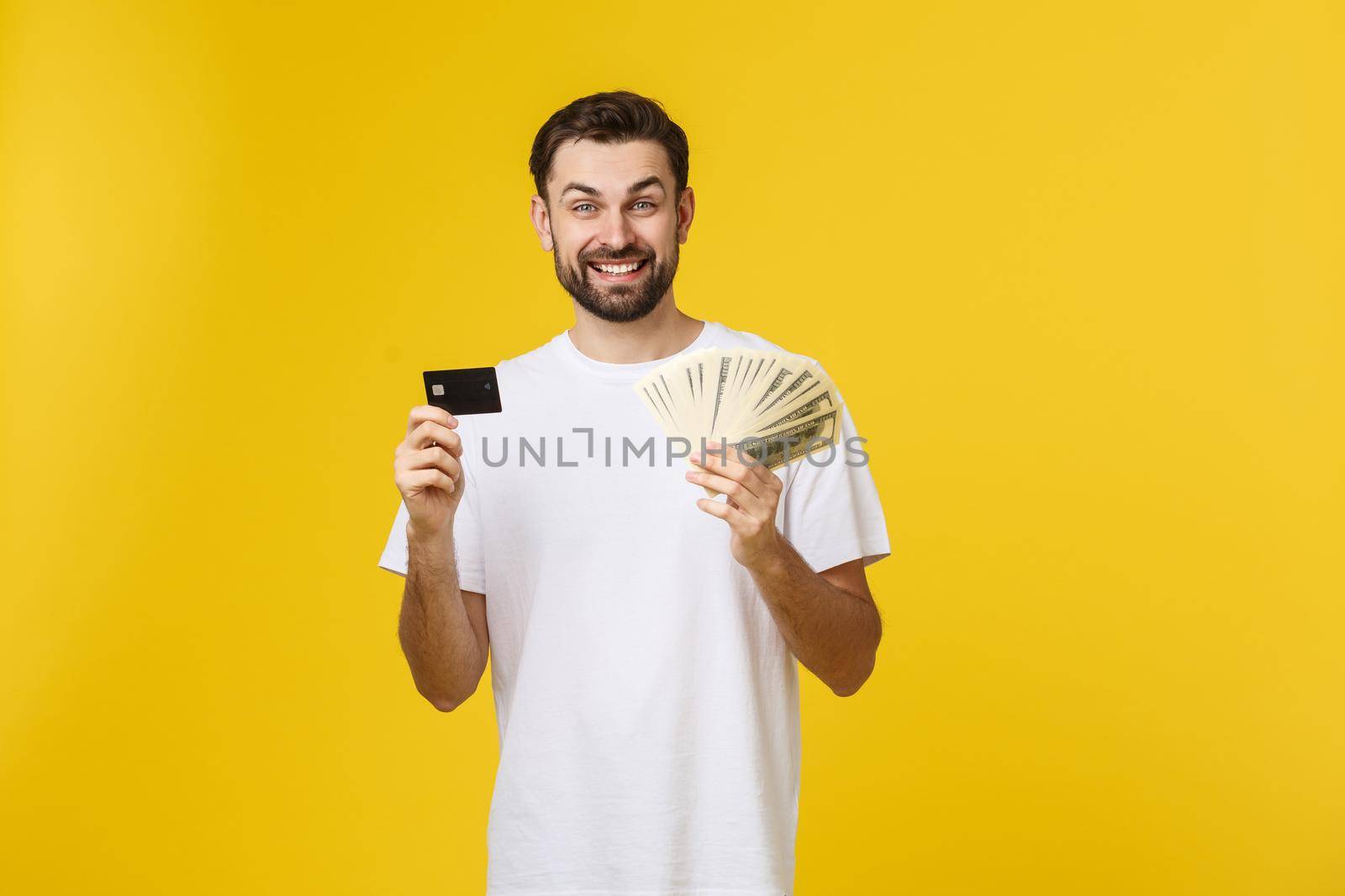 Young handsome happy smiling man holding banking card and cash in his hands isolated on yellow background