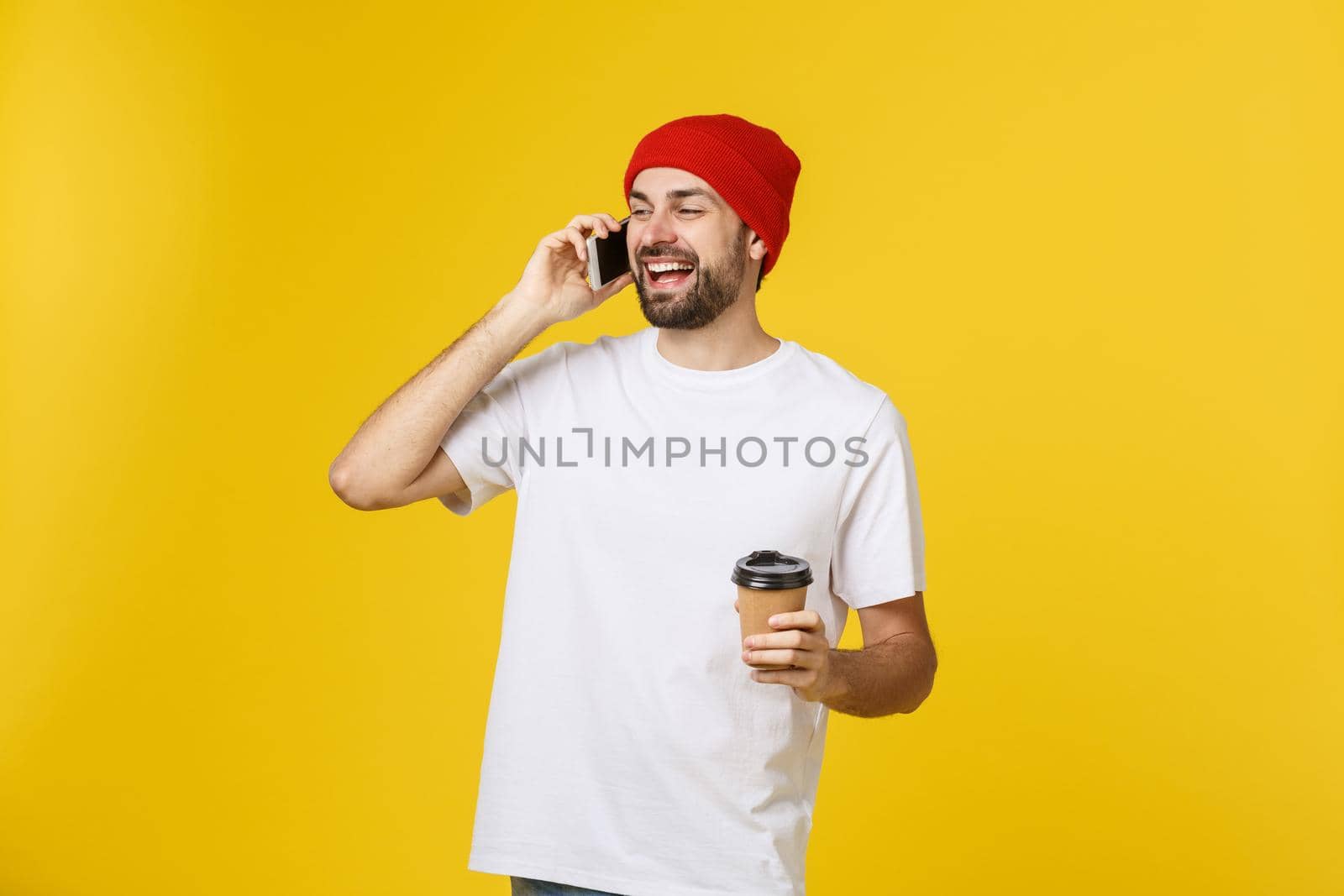 Portrait of a cheerful young man wearing casual clothes standing isolated over yellow background, holding mobile phone, drinking takeaway coffee. by Benzoix