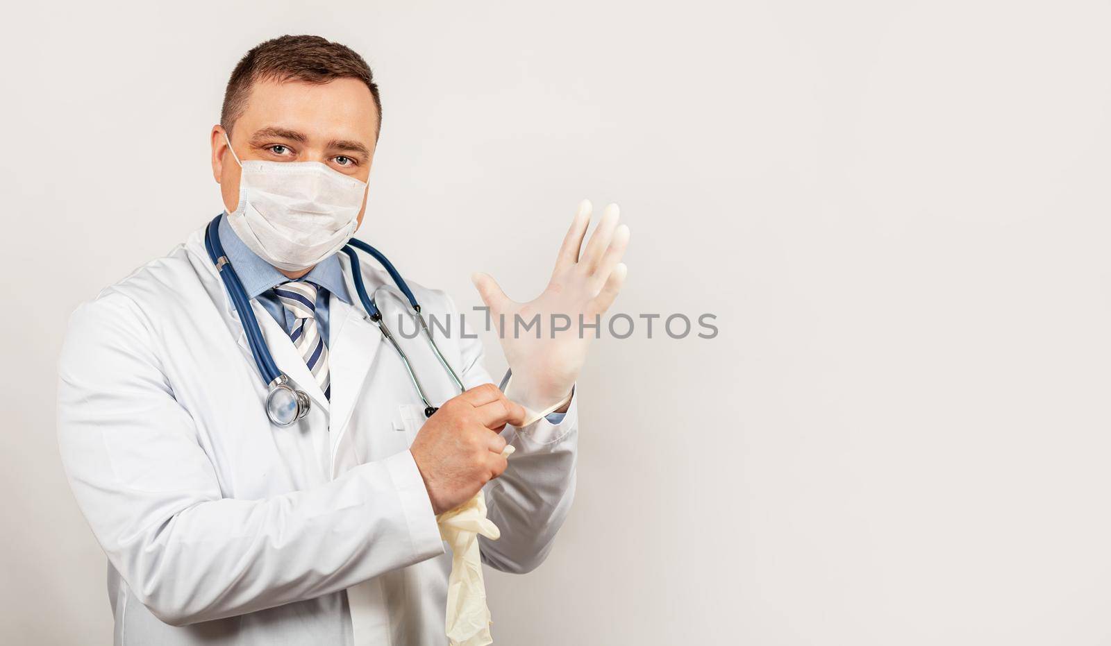 Portrait of a friendly doctor in medical mask and lab coat putting on sterile gloves and looking at the camera on a white background