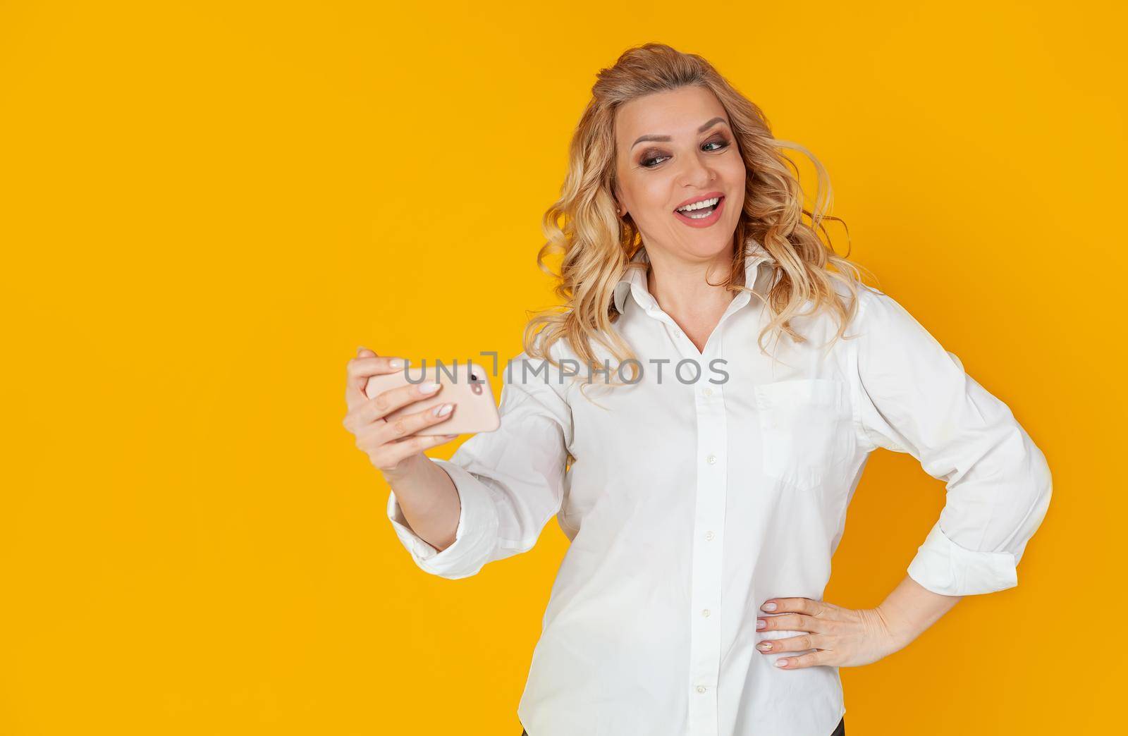 Portrait of a stylish beautiful smiling caucasian blonde woman taking a selfie on a smartphone, holding a mobile phone in outstretched hand and trying to look beautiful in the photo, yellow background