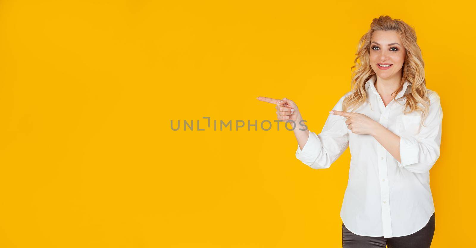 Lovely enthusiastic cute blond woman in a white shirt, pointing fingers to the left and smiling, excitedly shows her friend a terrific new place, presents copy space, hurries to try the product